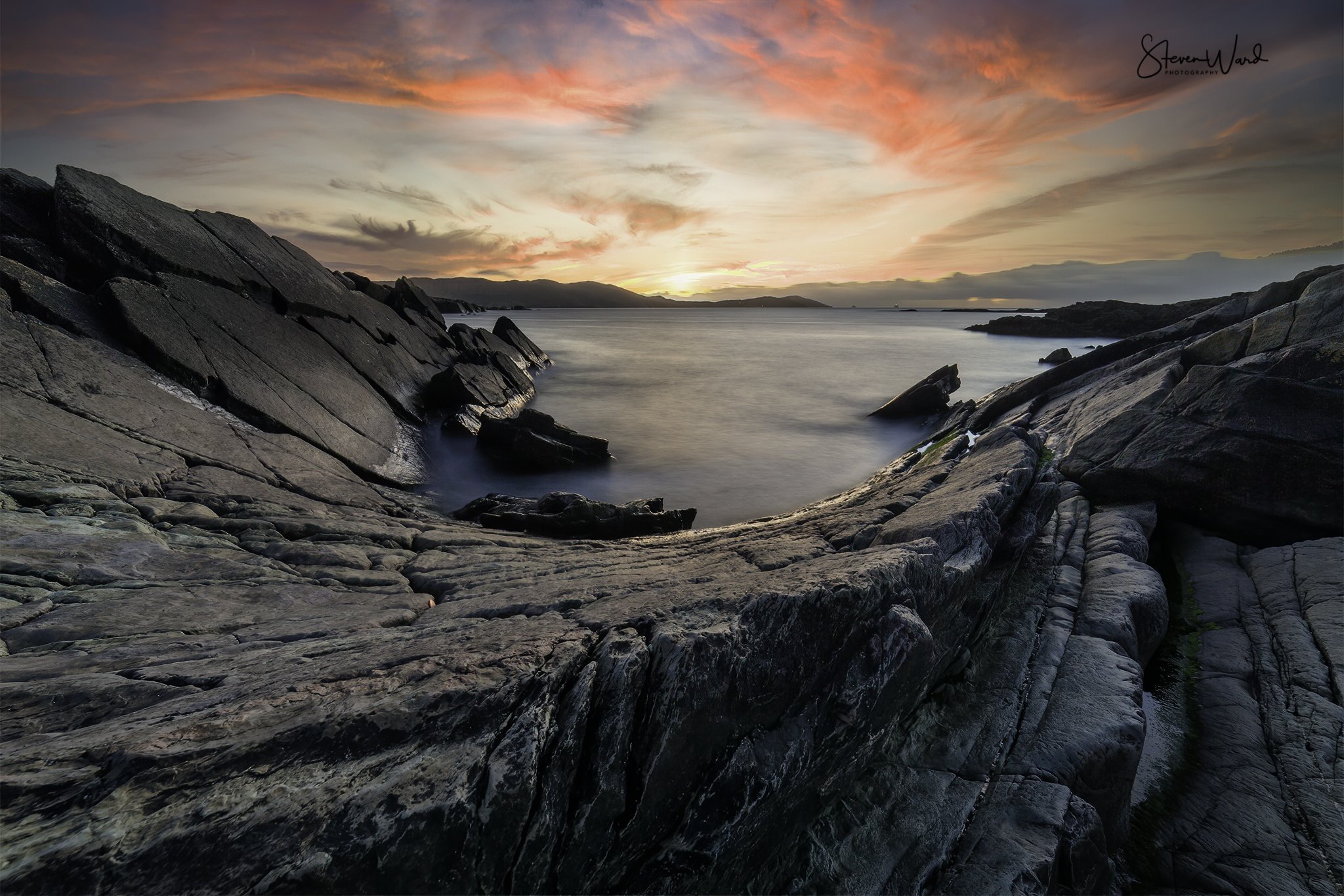 Sunset over rocky coastline and calm water with mountains in the background.