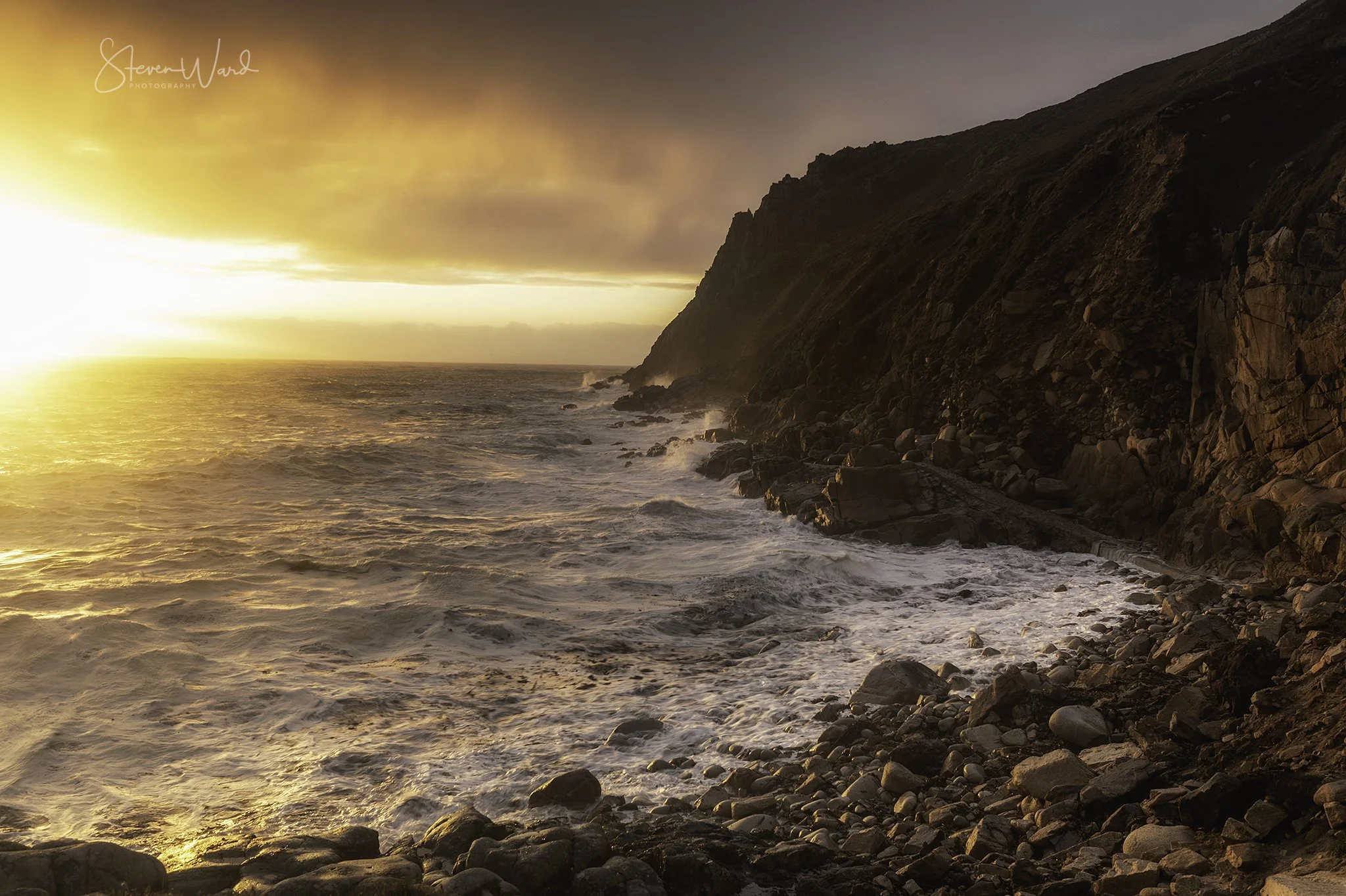Sunset over a rocky coastal shoreline with waves crashing against the rocks and a cliff on the right side.