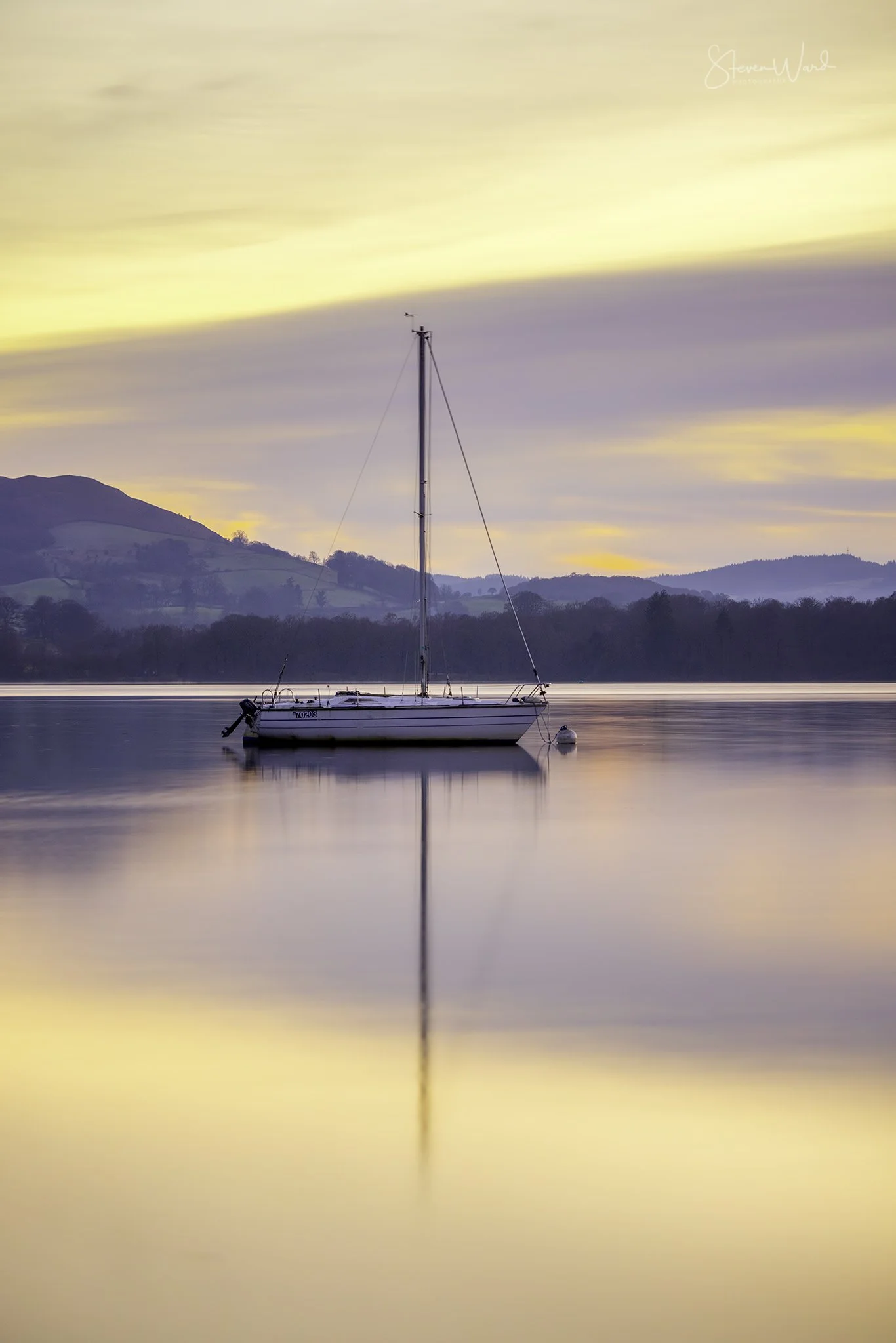 A sailboat floating on calm water during sunset with reflections and distant hills in the background.