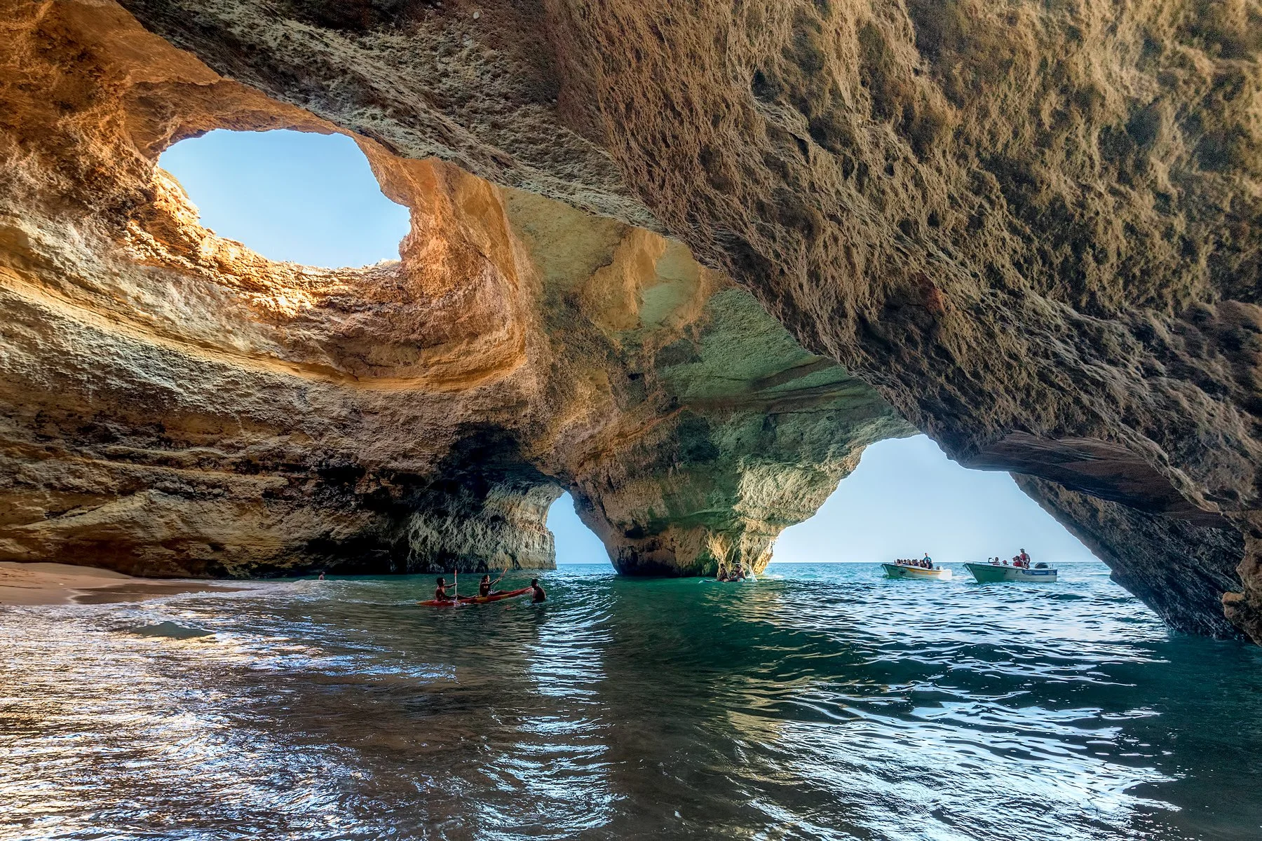 People kayaking and boating near large rock formations and caves on a sunny day with clear blue sky.
