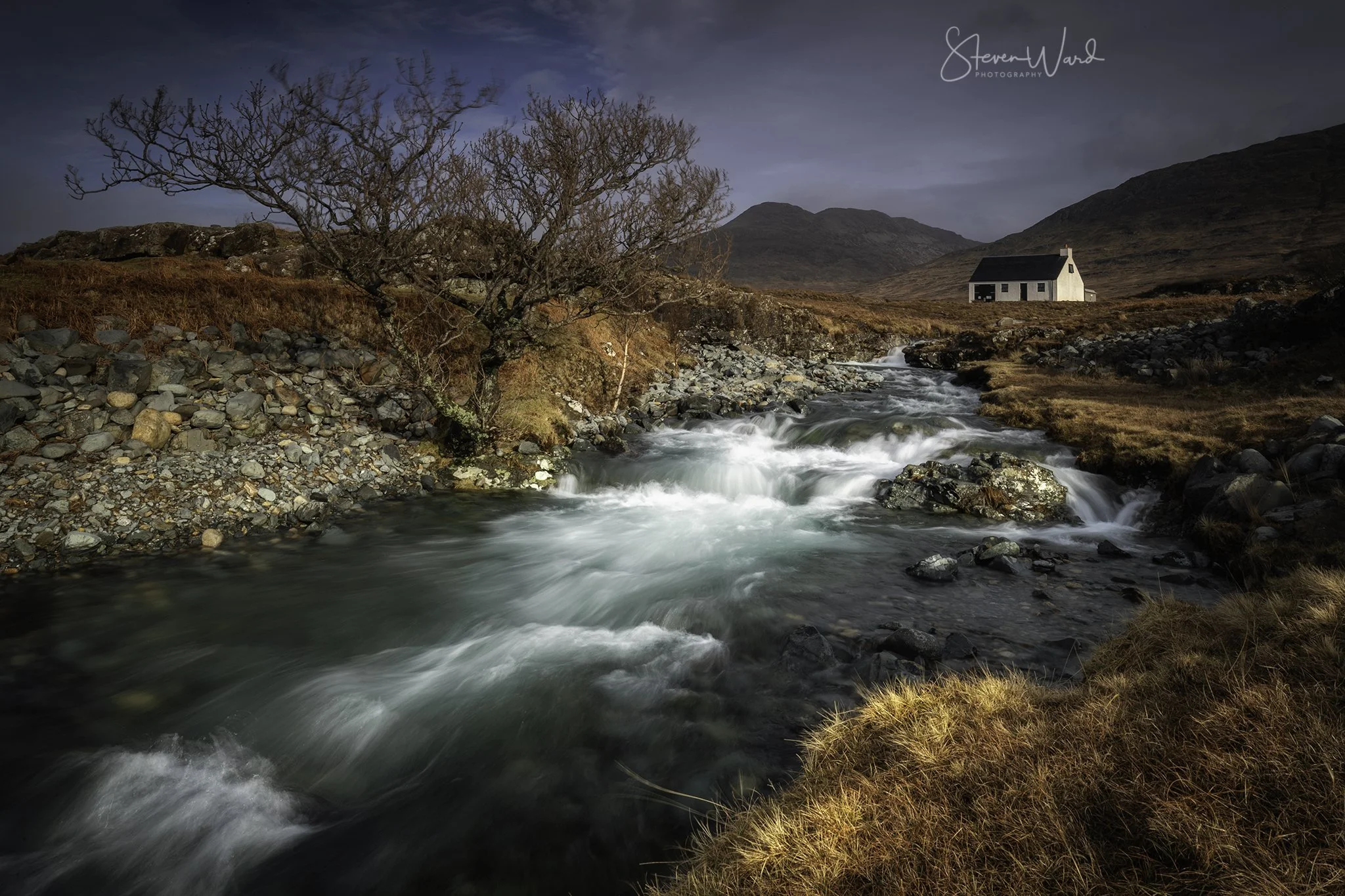 A scenic landscape of a flowing river with rocks on its banks, a bare tree on the left, and a small white house in the distance, with mountains in the background under a cloudy sky.