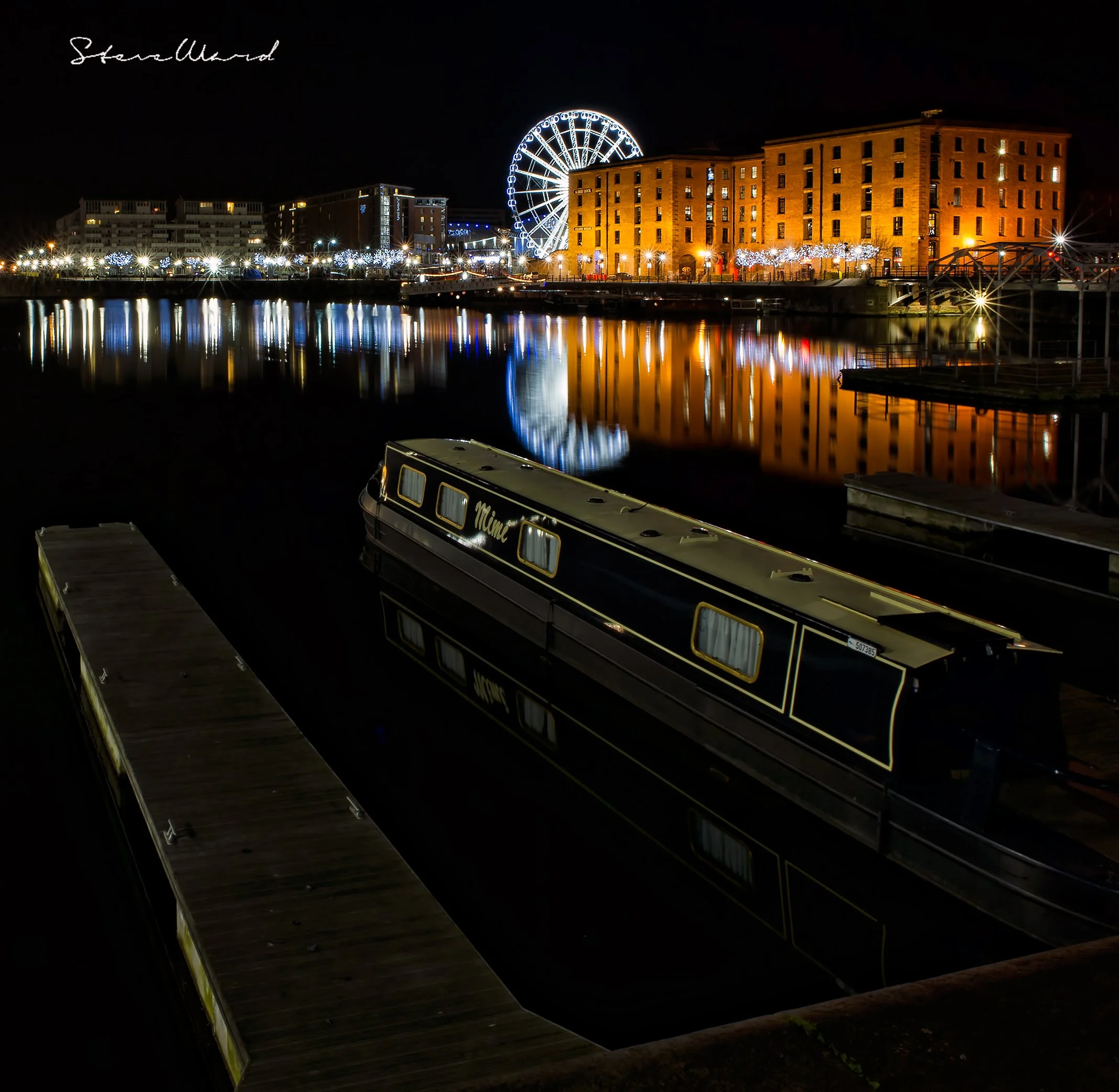 Night view of a harbor with a bridge and a Ferris wheel illuminated with white lights, reflecting on the water, alongside a large brick building. A boat named 'Miami' is docked in the foreground.