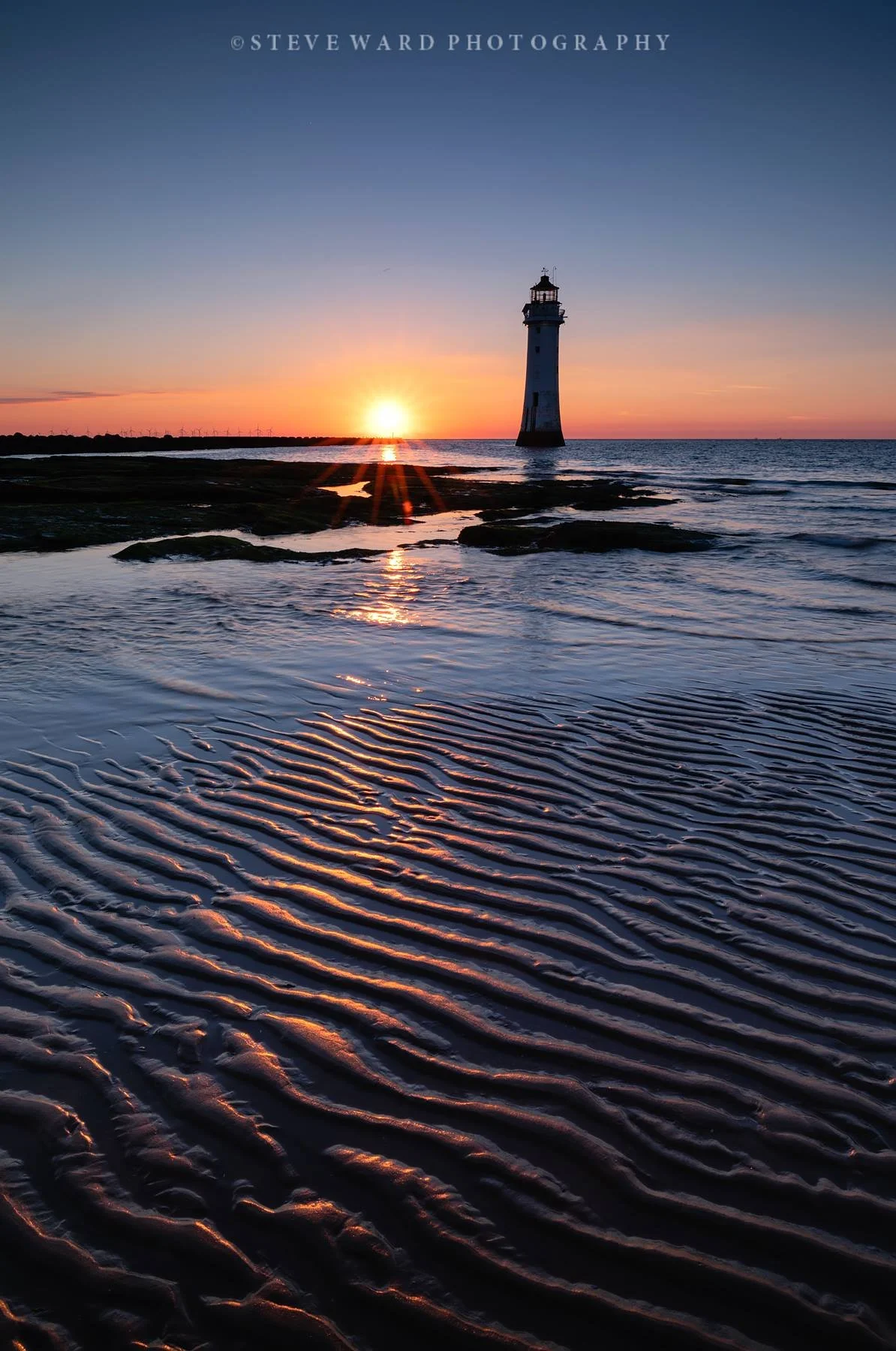 A lighthouse on a beach at sunset, with the sun near the horizon and its reflection in the water showing rippled sand patterns.