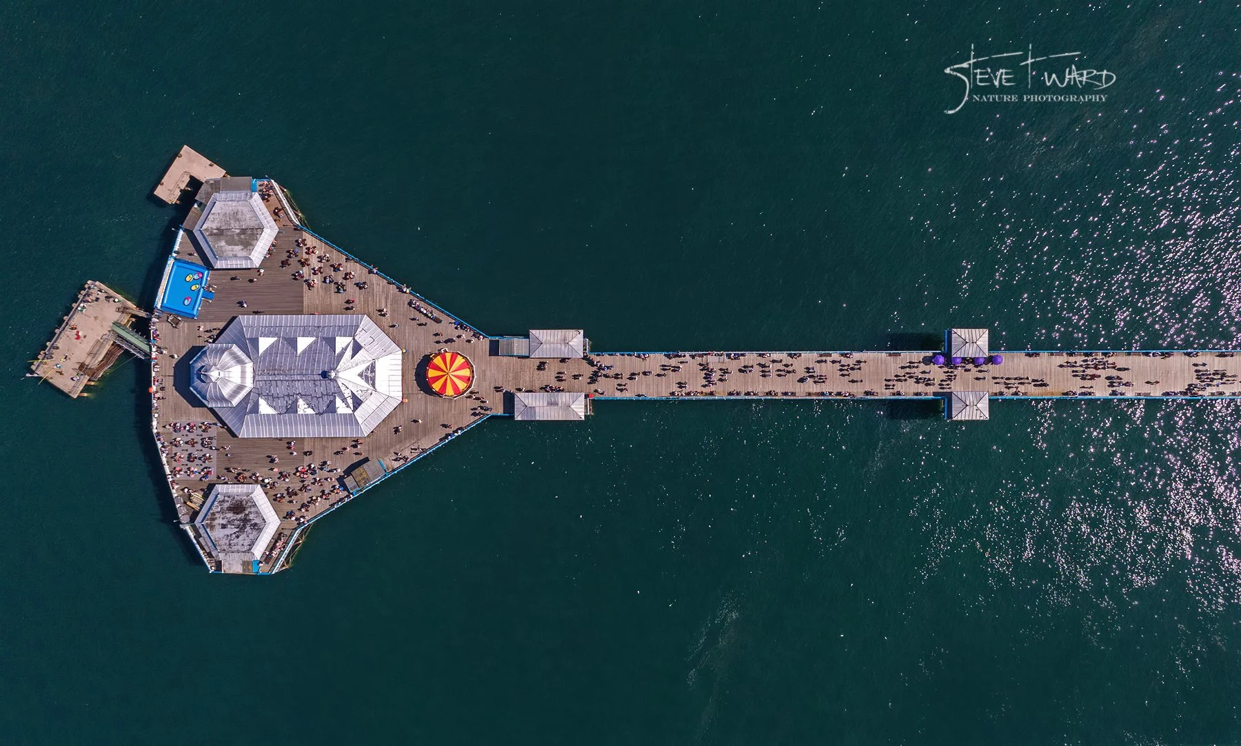 Aerial view of a pier extending into a body of water with a crowd of people walking on it and some gathering around structures at its end.