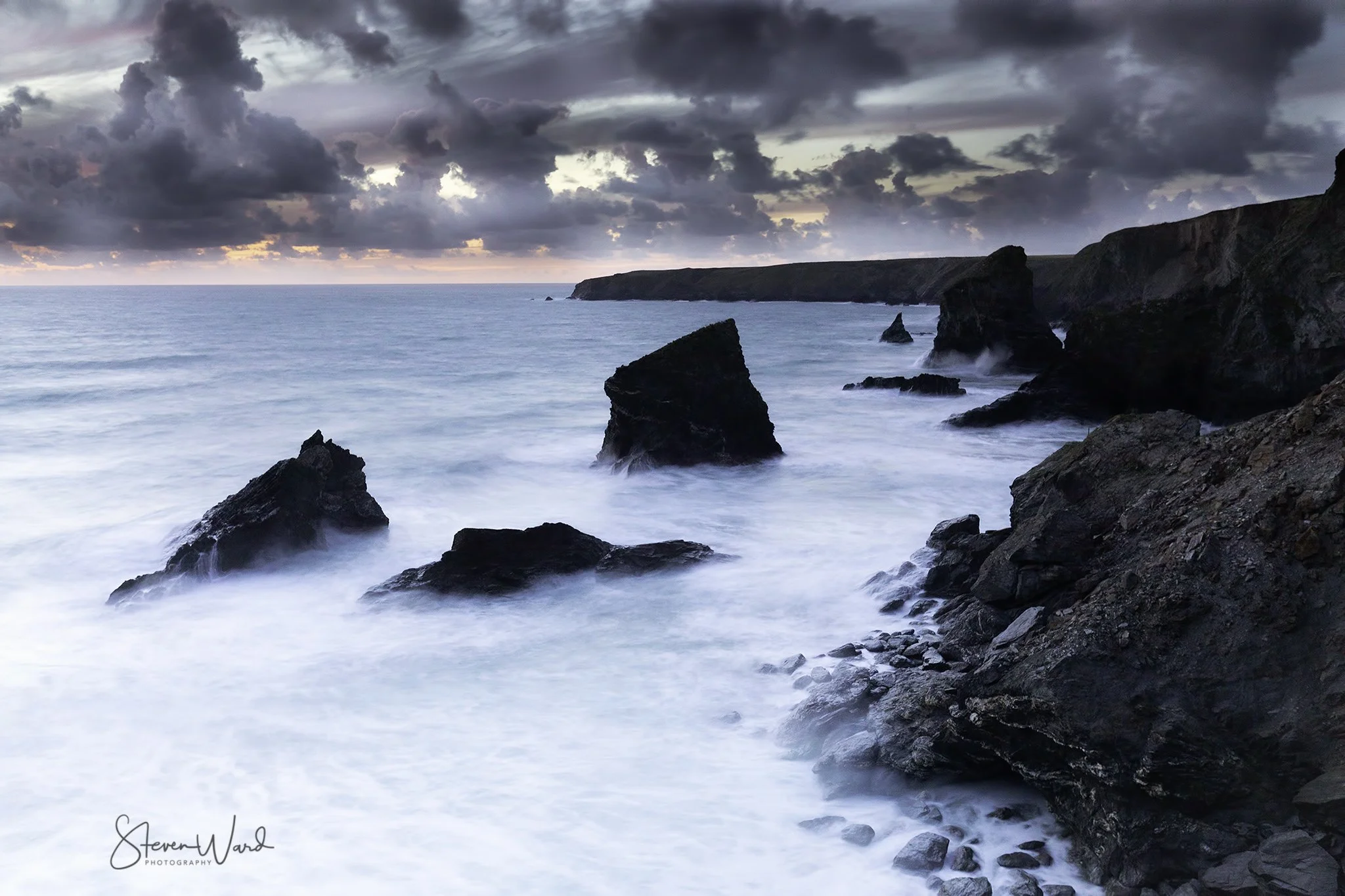 A rocky coastline at sunset with dark clouds overhead and waves crashing against large rocks in the ocean.