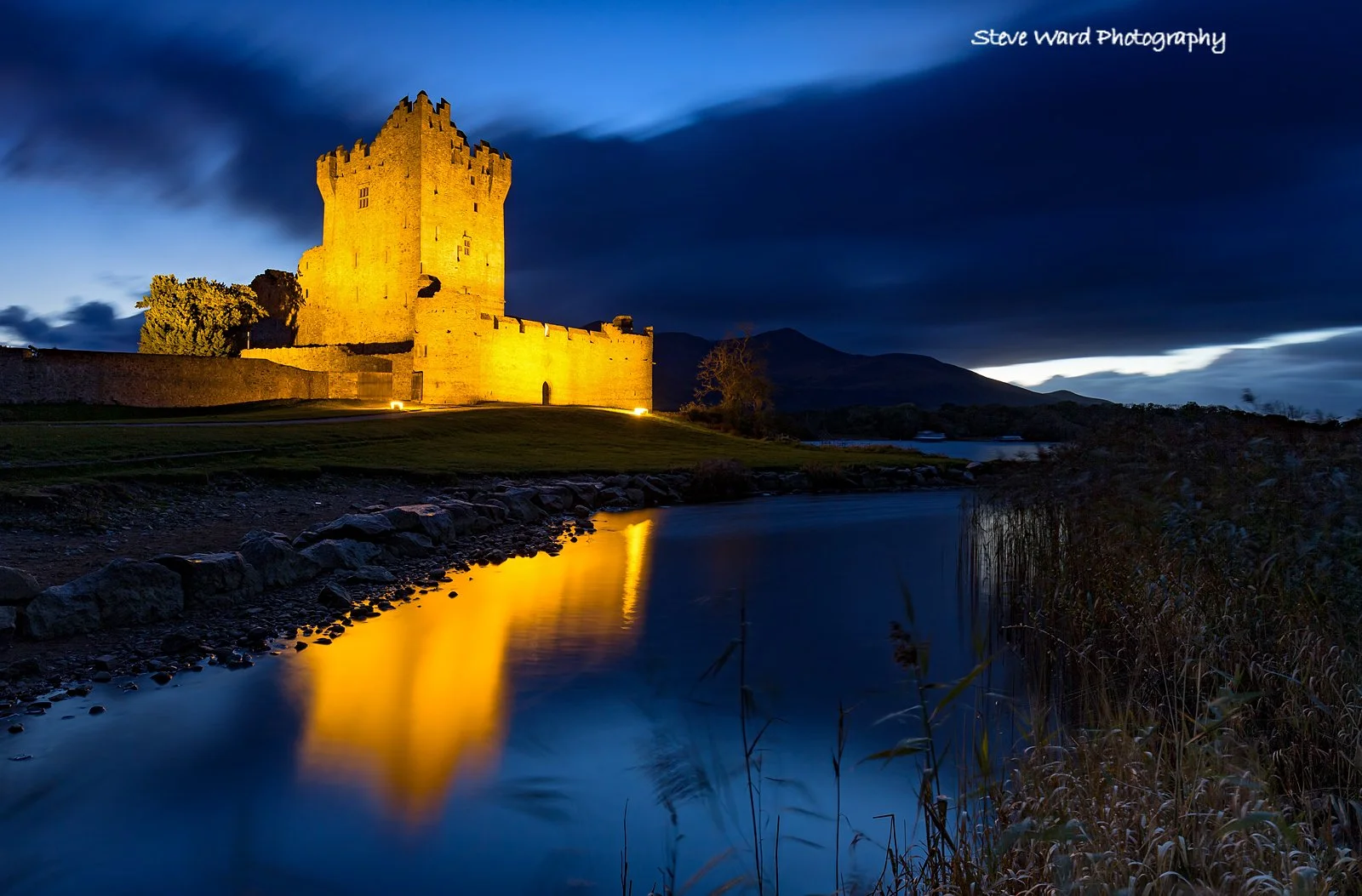 A castle illuminated at night with yellow lighting, beside a calm river reflecting the structure. Dark clouds and mountains are visible in the background.