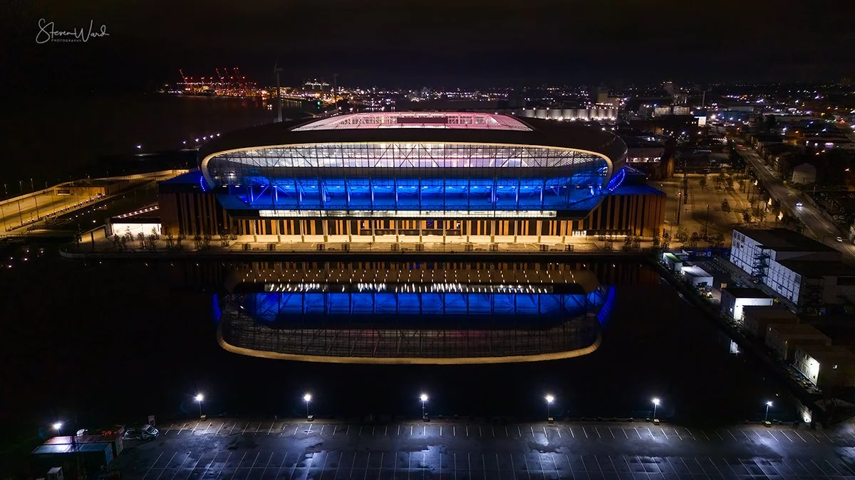 Night view of a large, modern stadium lit in blue and pink, with its reflection visible in a body of water in front, surrounded by an empty parking lot.