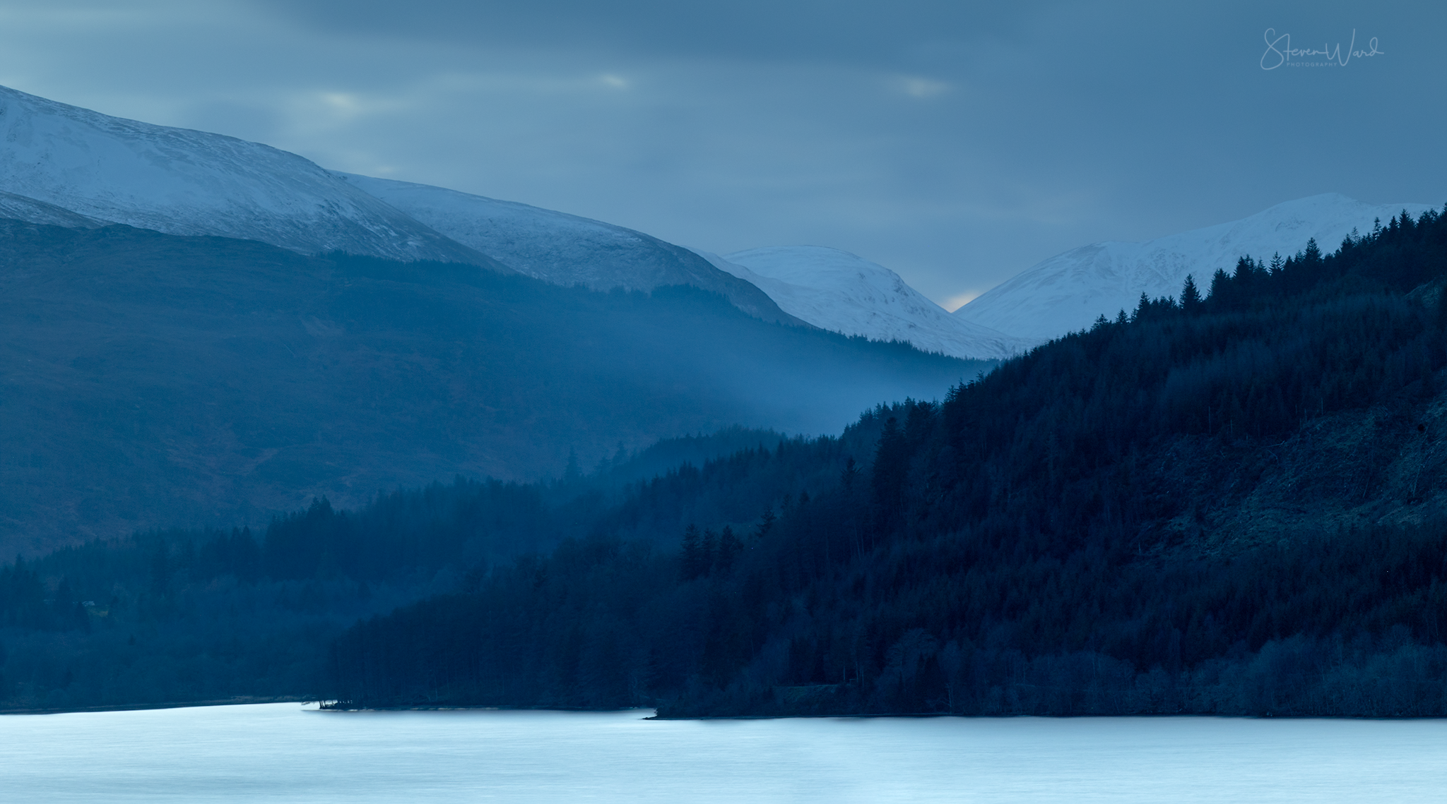A scenic view of a mountain landscape with snow-capped mountains in the background, dense forested hills in the middle ground, and a calm body of water in the foreground, all under a cloudy sky.