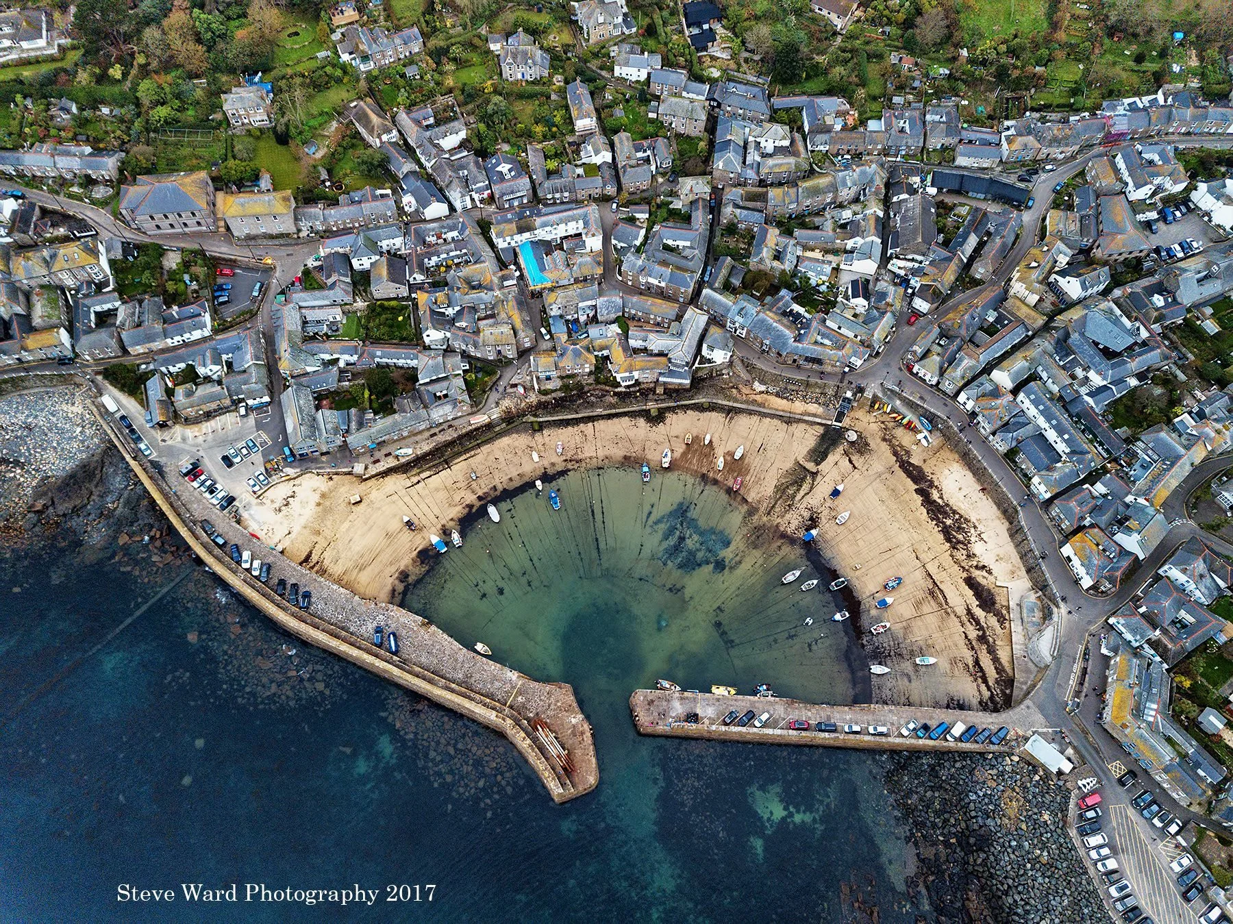An aerial view of a small coastal town showing tightly packed houses, narrow streets, and a harbor with boats docked on a sandy shore, with surrounding greenery and rocky shoreline.