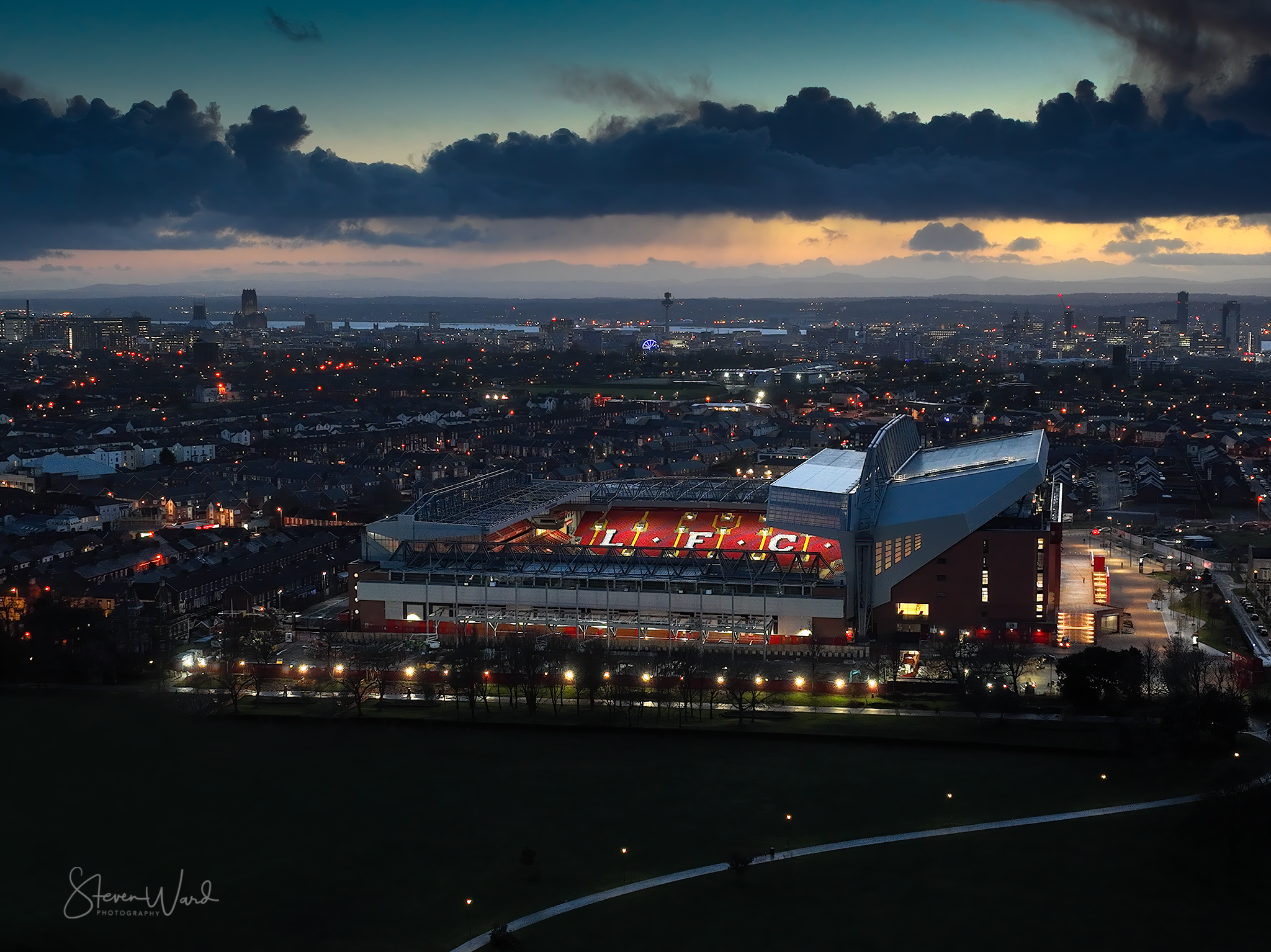 Aerial view of a city at dusk featuring Old Trafford football stadium illuminated and in the foreground, with a skyline of buildings and a partly cloudy sky in the background.