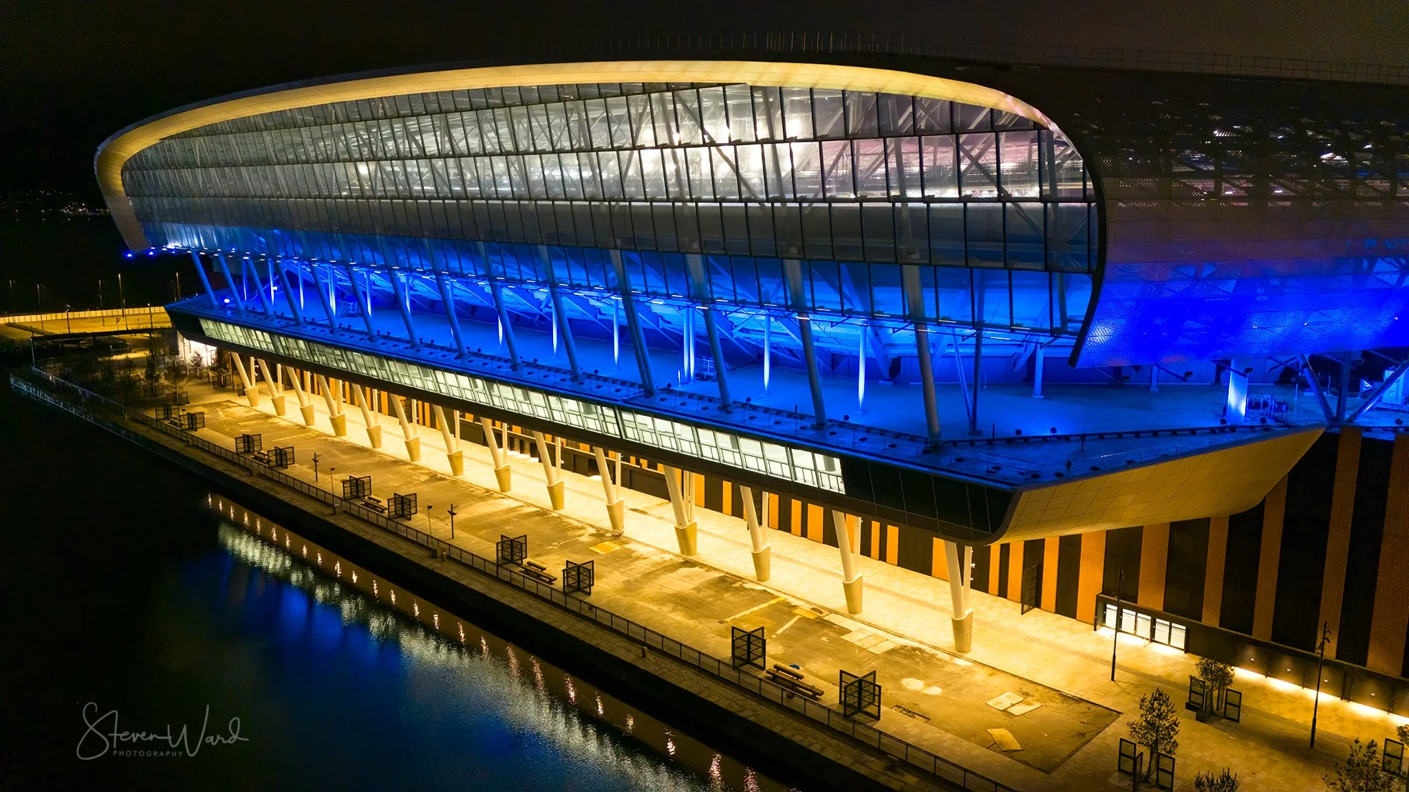 A modern, architecturally distinctive building illuminated with blue and yellow lights at night, situated along a body of water, with benches and streetlights on the promenade.
