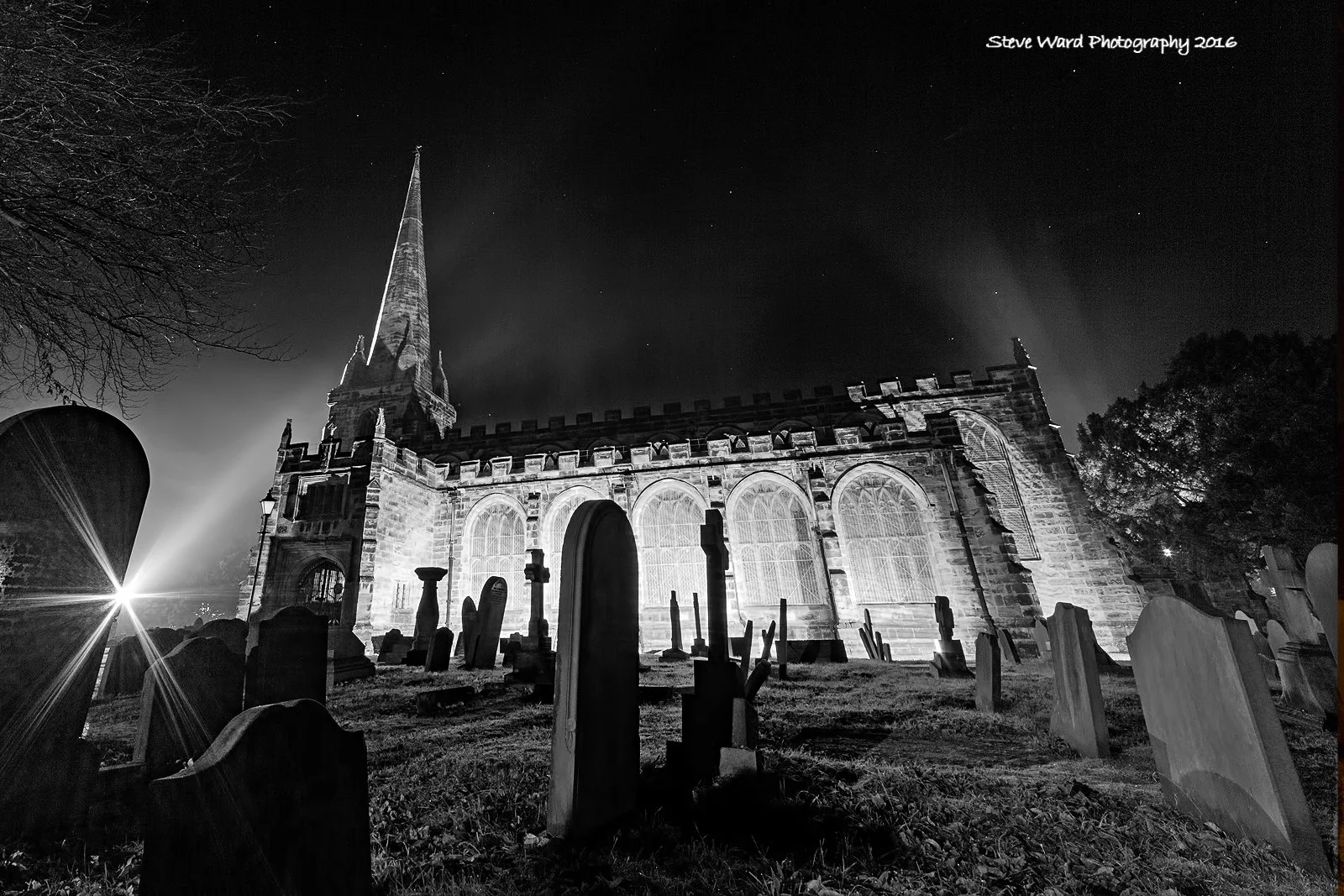 Black and white night photo of an old church with a tall steeple, illuminated from the front, with a graveyard in the foreground and a star in the sky.