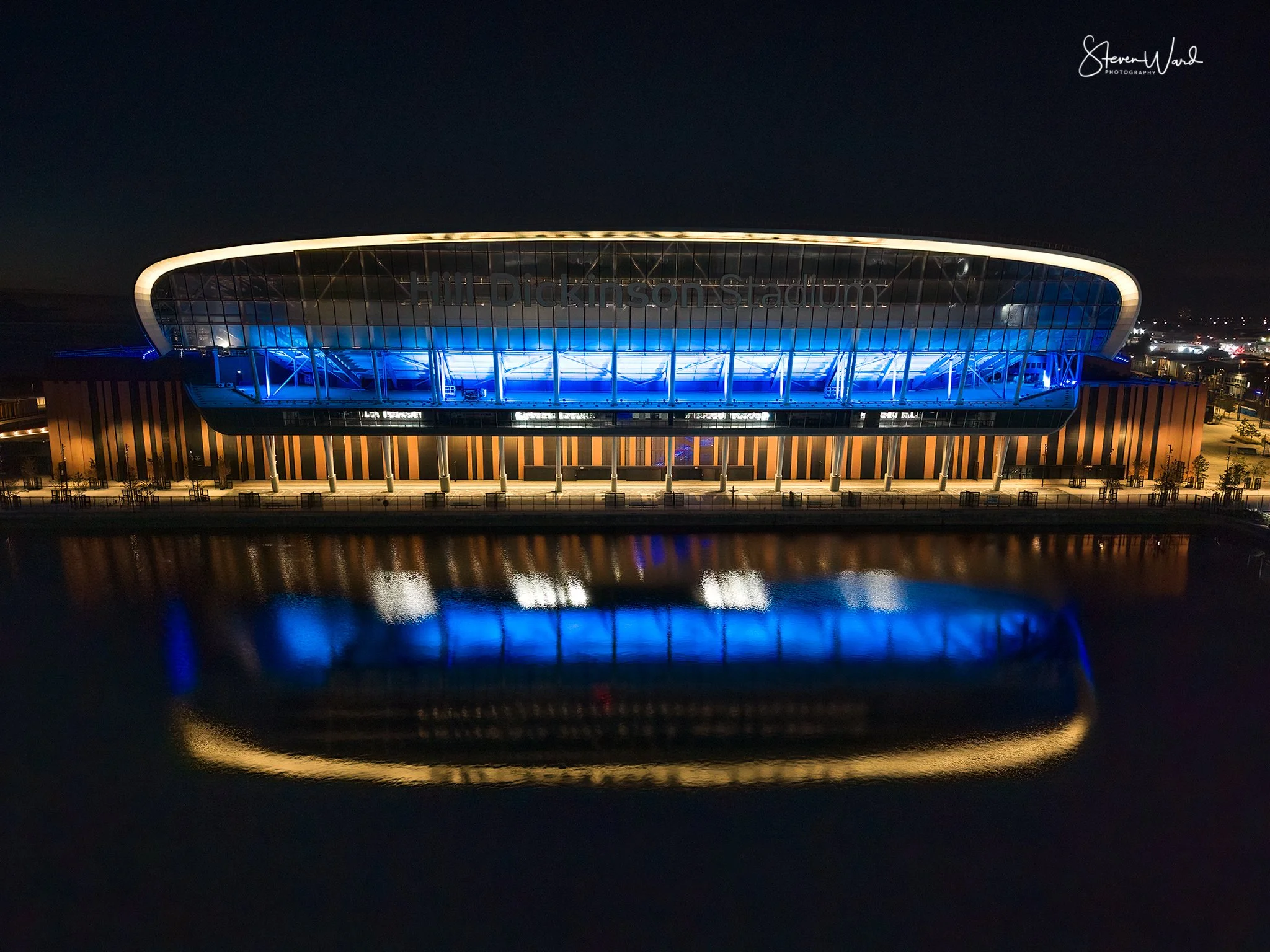 Nighttime view of Field Dickenson Stadium with blue lighting, reflection visible on the water in front, and building's name partially illuminated, with a dark sky and city lights in the background.