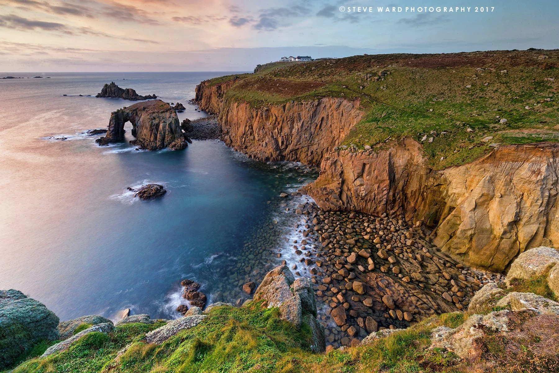 A scenic view of a rocky coastal landscape with a natural rock arch formation in the water, surrounded by cliffs, green vegetation, and a small pebble beach, at sunset.