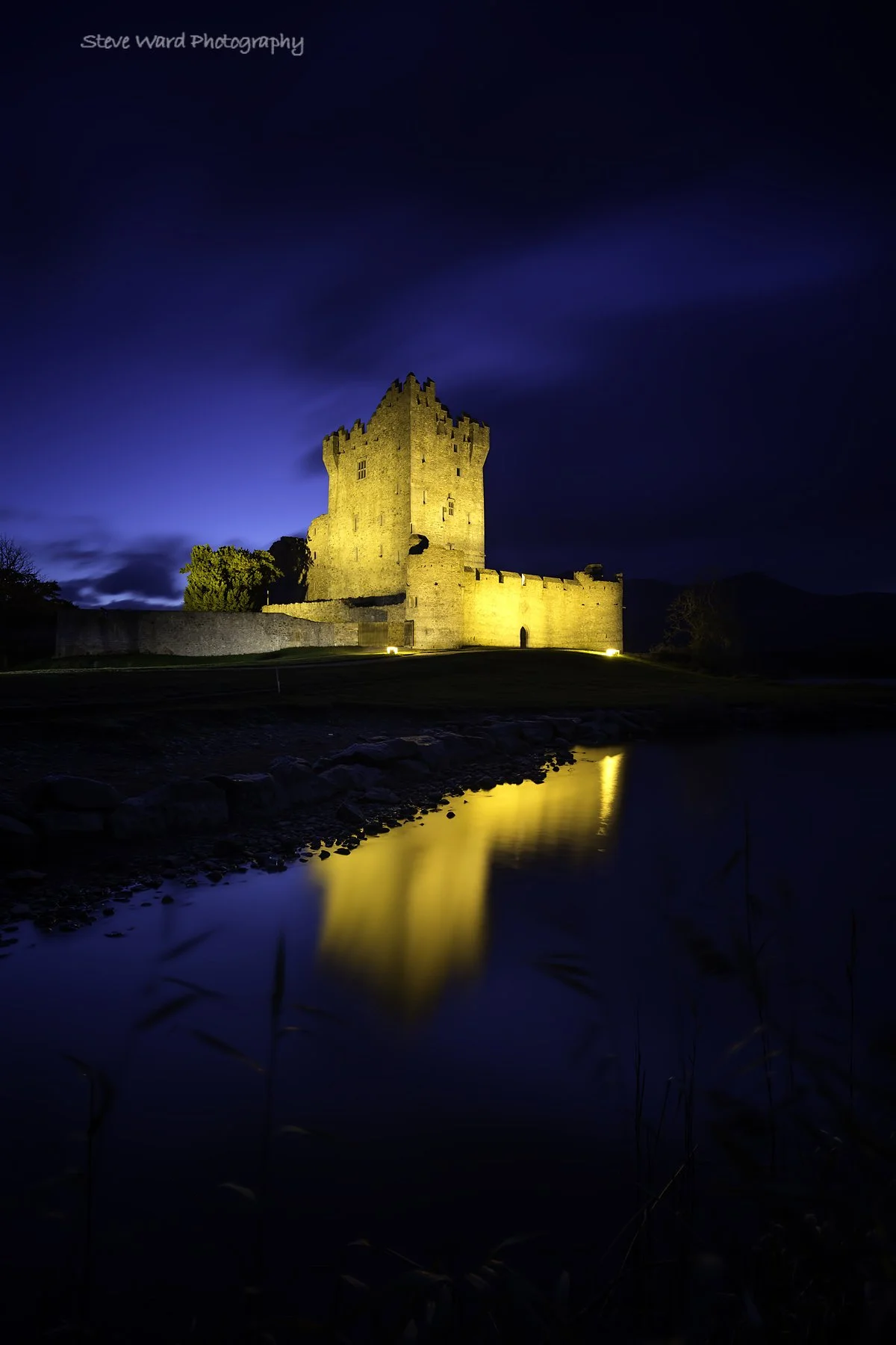 A historic castle illuminated at night with its reflection seen in a calm body of water in the foreground.