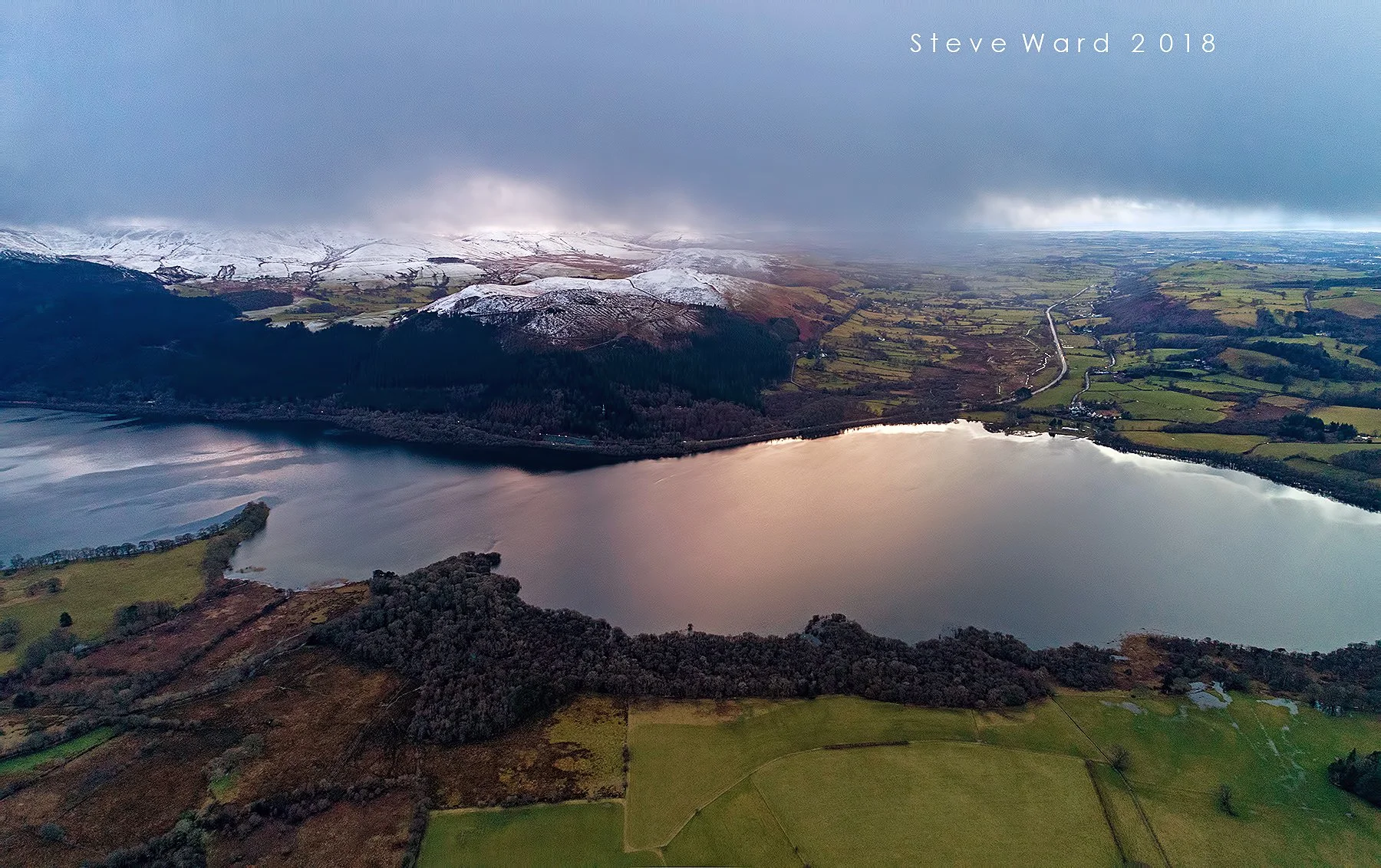 Aerial view of a lake surrounded by green fields, dark forests, and snow-capped mountains under a cloudy sky.
