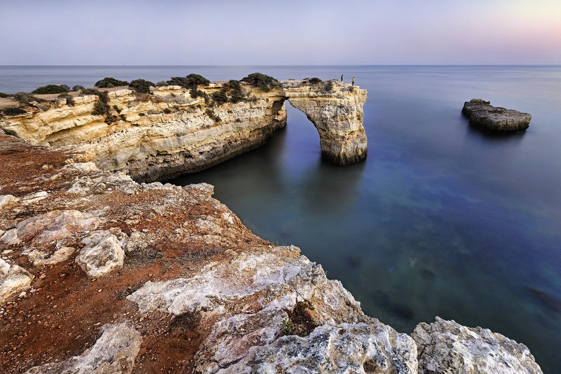Cliffs with an arch formation by the ocean, with two people standing on the edge, a large rock in the water nearby, and a calm sea in the background during sunset or sunrise.