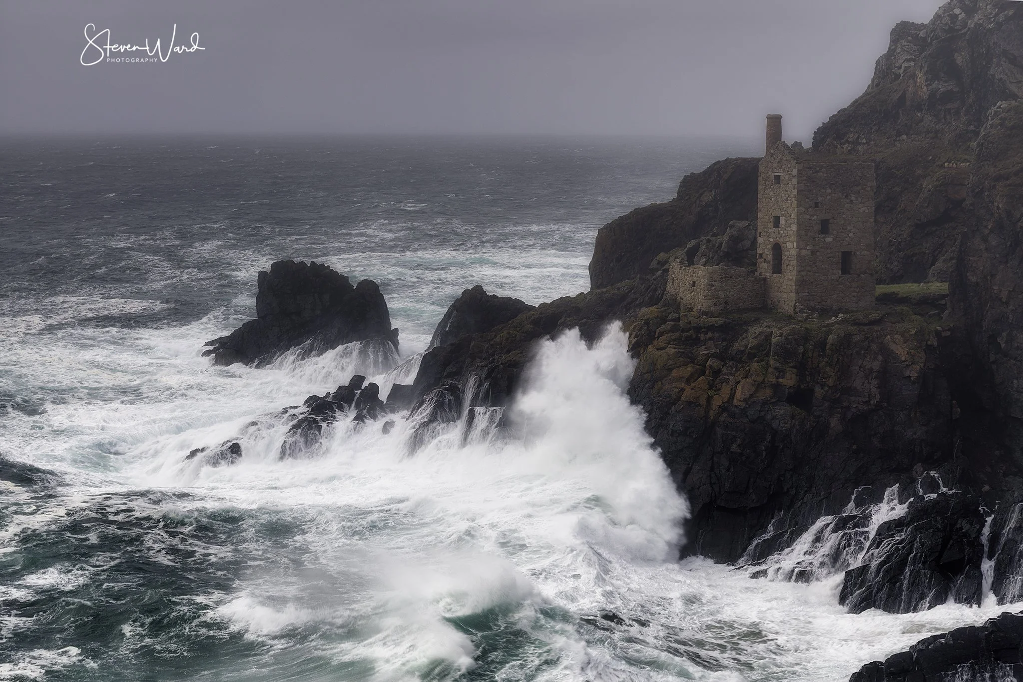 A stormy ocean coast with waves crashing against dark rocks and an old stone building on the cliffside.