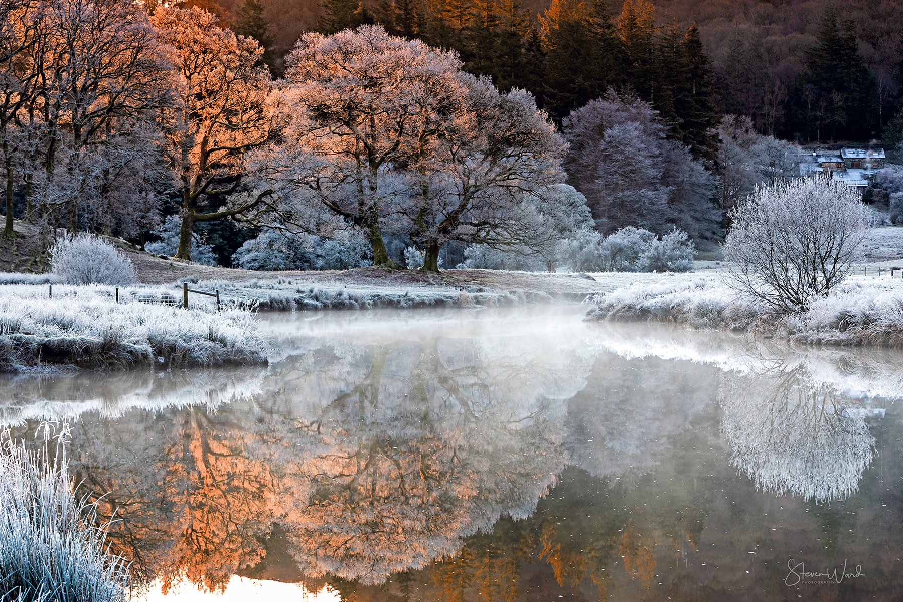 A winter scene of a river with icy, snow-covered banks and frosted trees. The trees are backlit by early morning or late afternoon sunlight, creating a reflection in the water. There are houses on a hillside in the background.