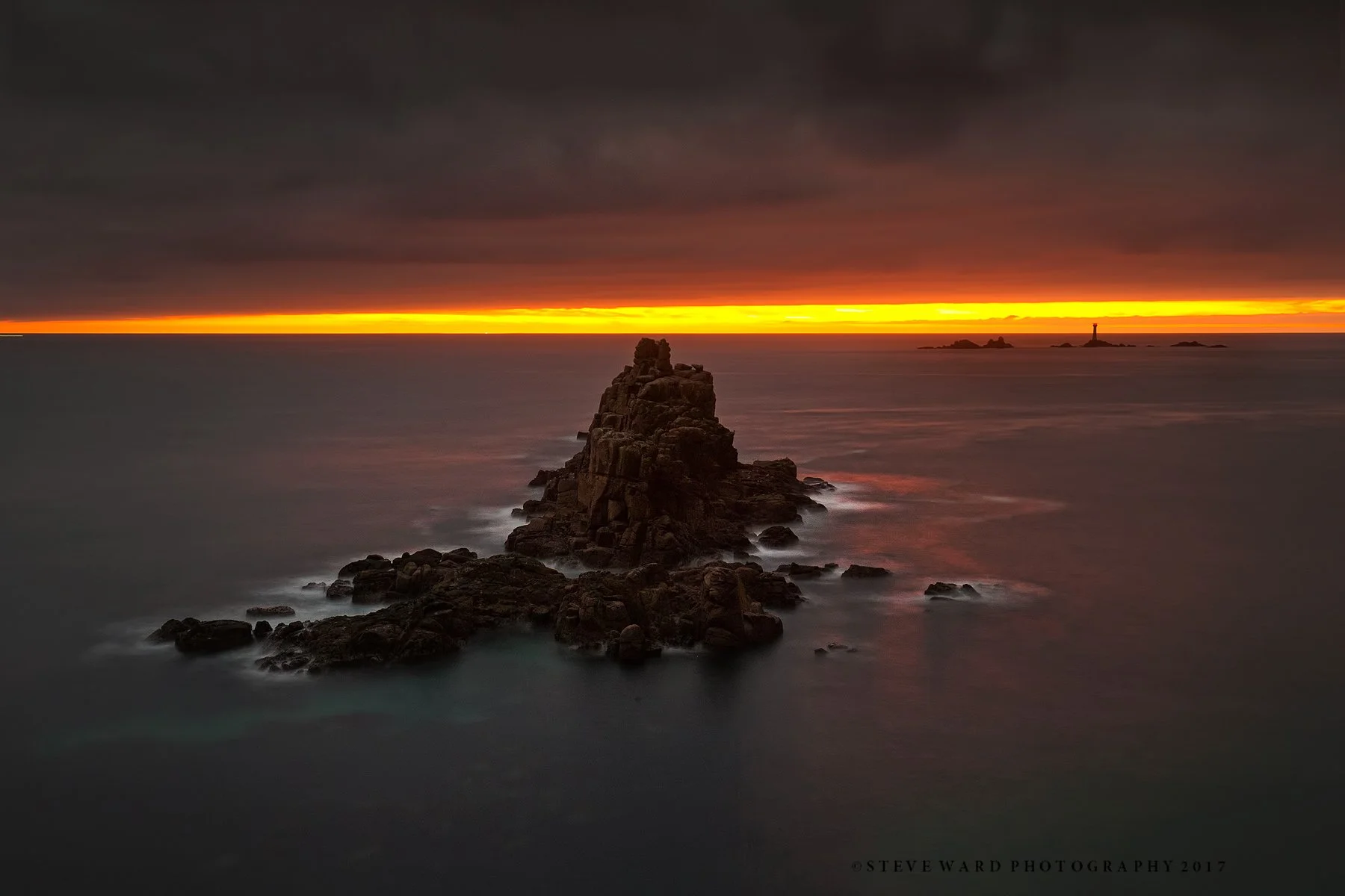 Long exposure photo of a rocky ocean cove during sunset with dark clouds and a vibrant orange and yellow horizon, lighthouse on distant rocks.
