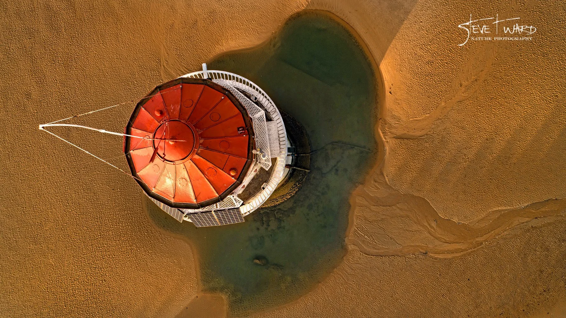 A bird's eye view of a lighthouse with a red, octagonal roof surrounded by sandy terrain and a small body of water.