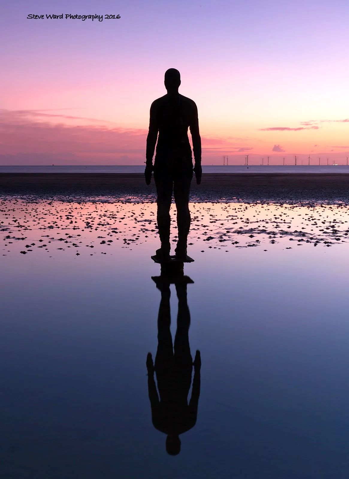 A person standing by shallow water on a beach during sunset with wind turbines visible in the background, their silhouette reflected on the water.