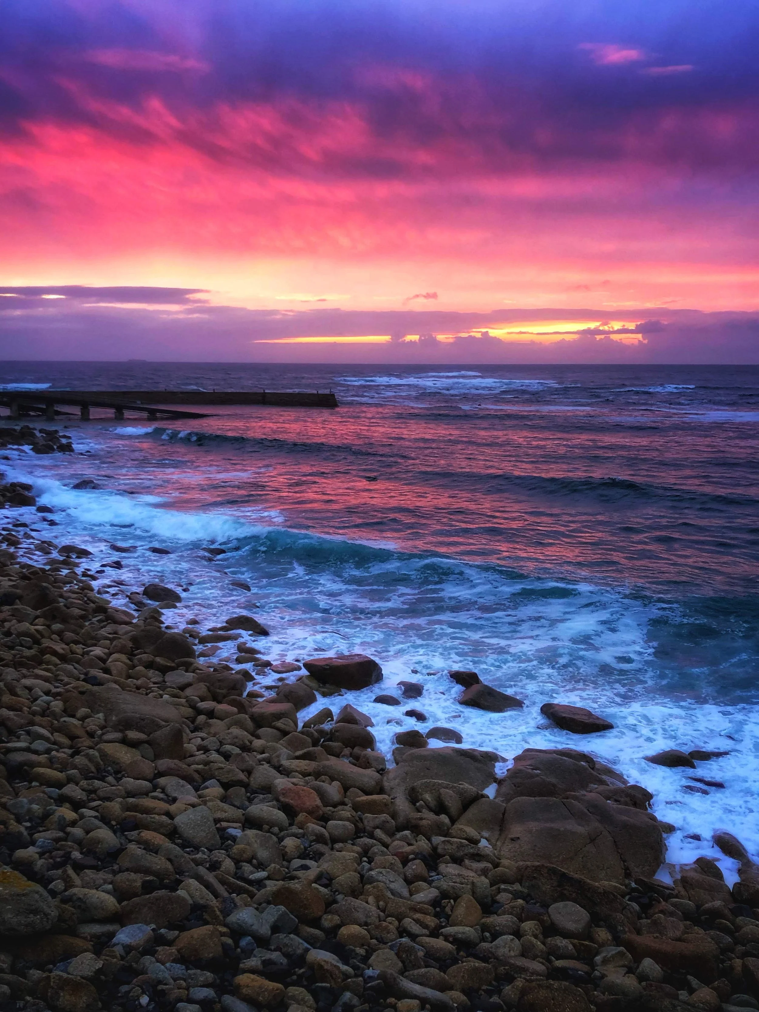 Colorful sunset over the ocean with pink and purple clouds, waves hitting a rocky shore, and a breakwater extending into the water.
