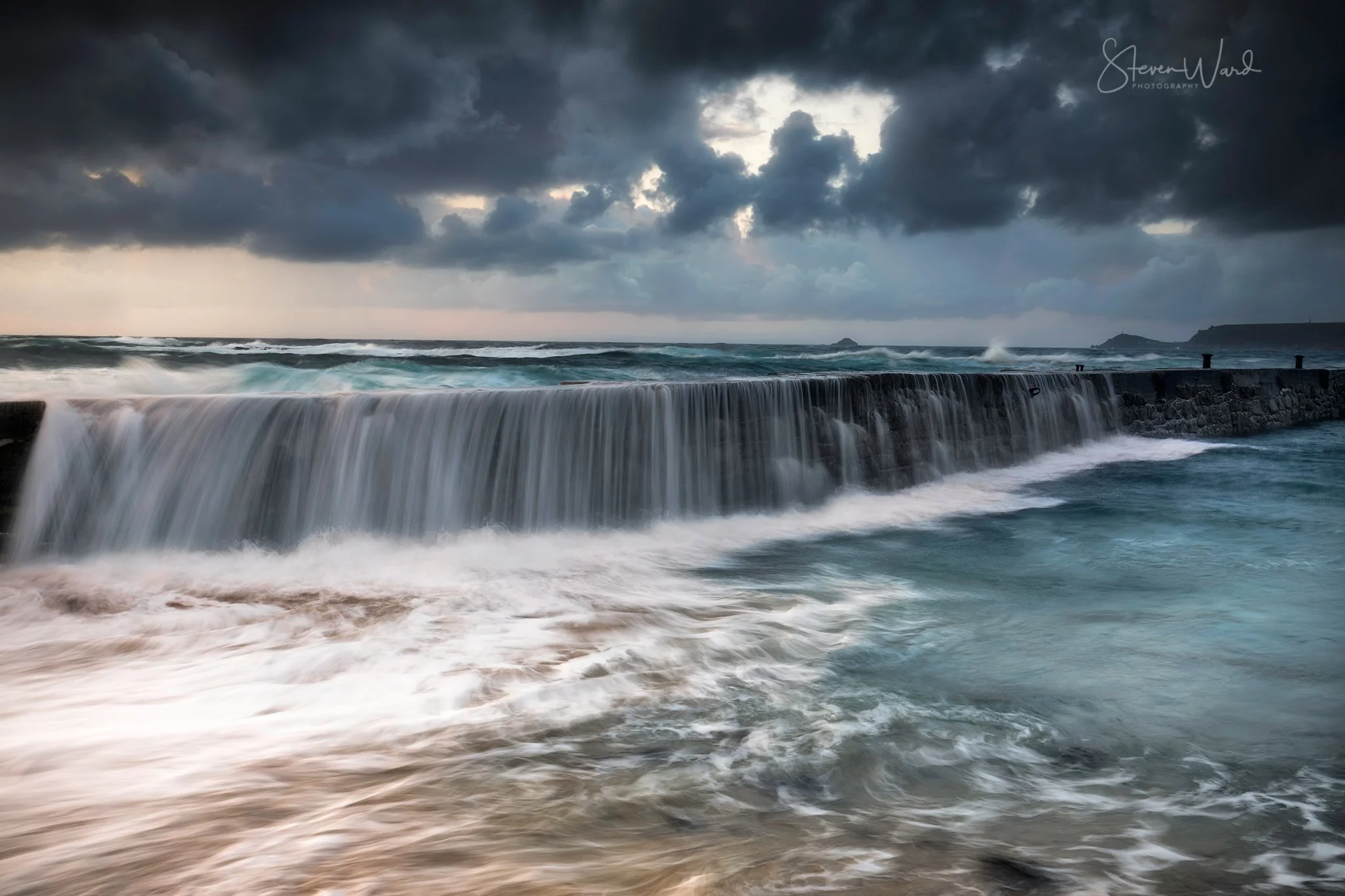 Overcast sky with dark clouds over a crashing ocean wave on a breakwater, with distant landforms on the horizon.