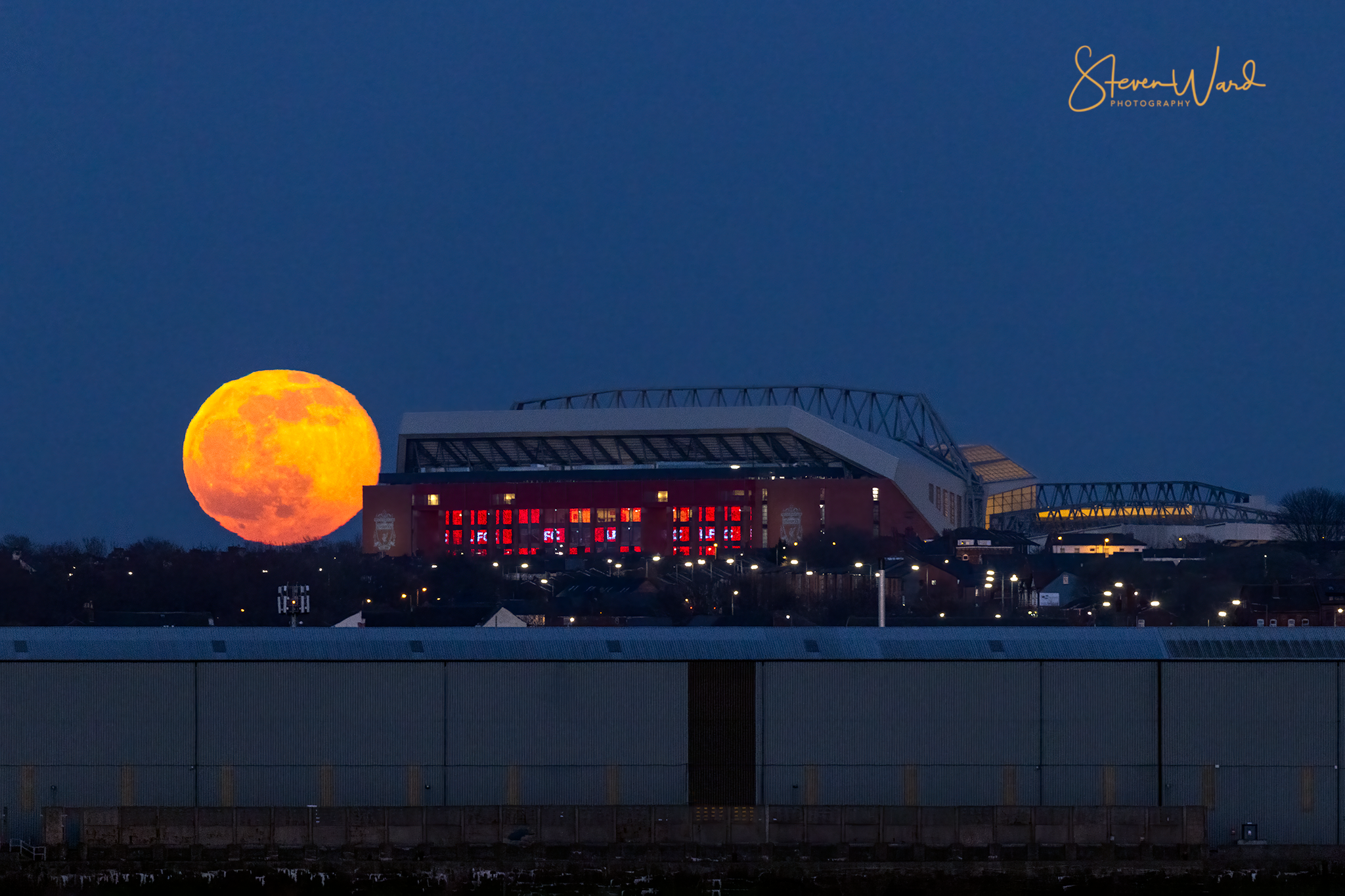 Full moon rising behind a football stadium at night with city lights in the foreground.