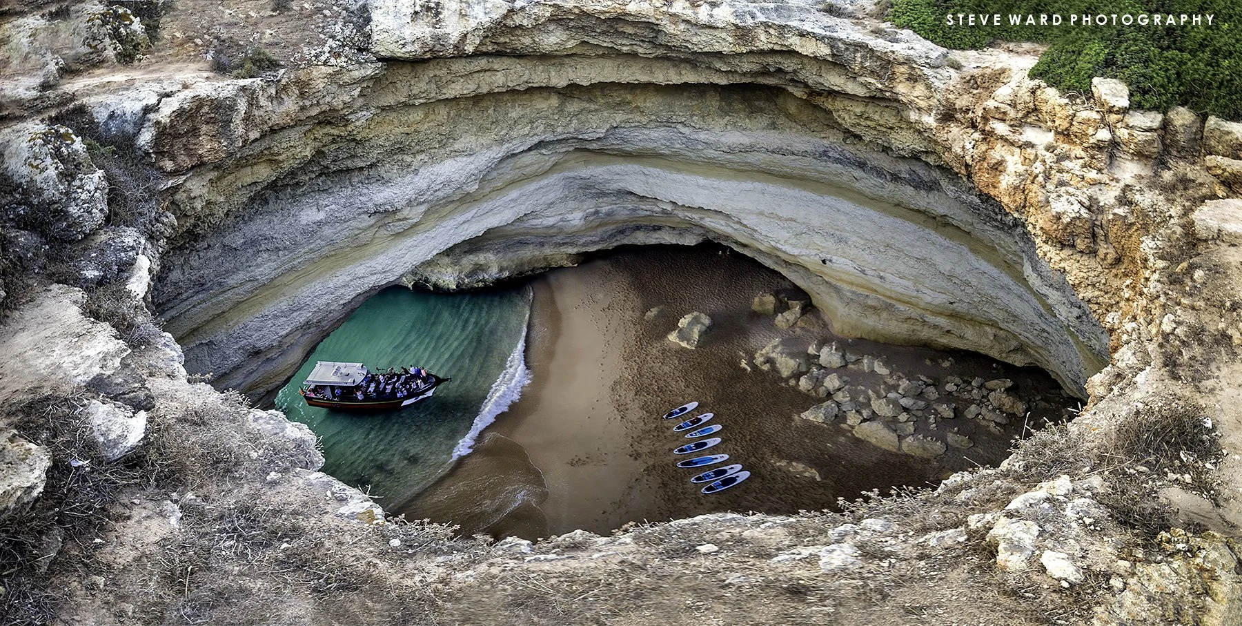 A boat with people on a turquoise and green water body inside a large circular natural rock formation with multiple layers of colorful sediment, overlooking a sandy beach with several paddleboards and rocks.