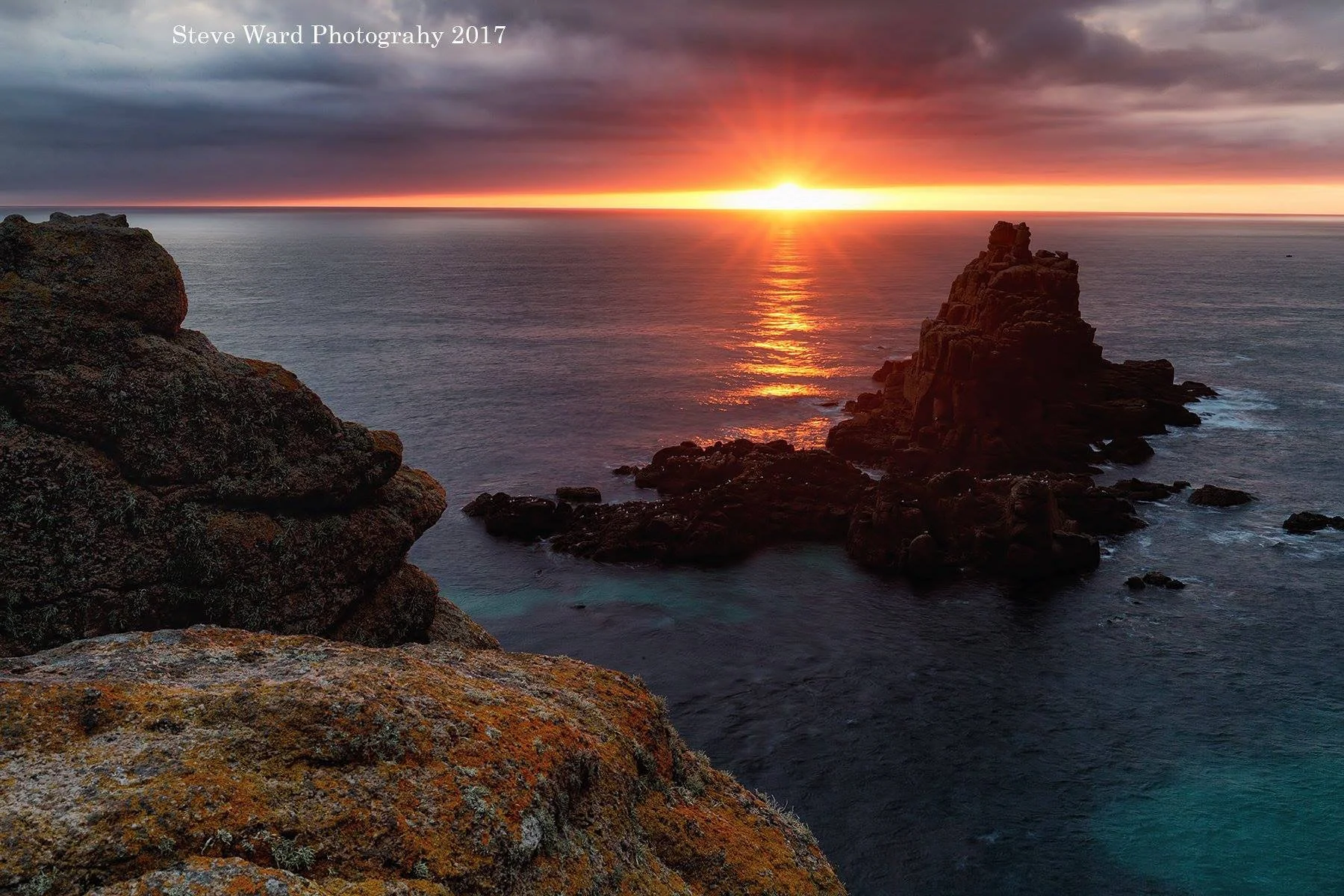 A scenic coastal view at sunset with rocky cliffs, a large rock formation emerging from the water, and the sun setting on the horizon, casting a warm glow across the sky and reflecting on the ocean surface.