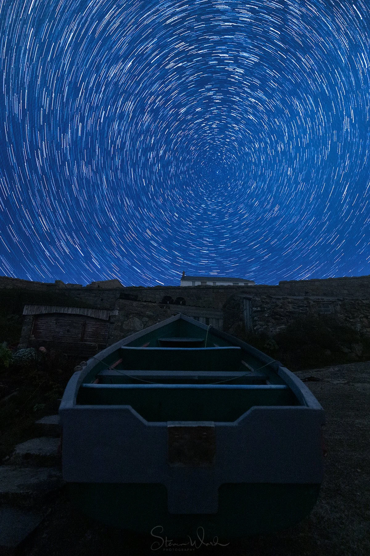 Long exposure night photograph of star trails in the sky with a boat in the foreground and a building on a hilltop in the distance.