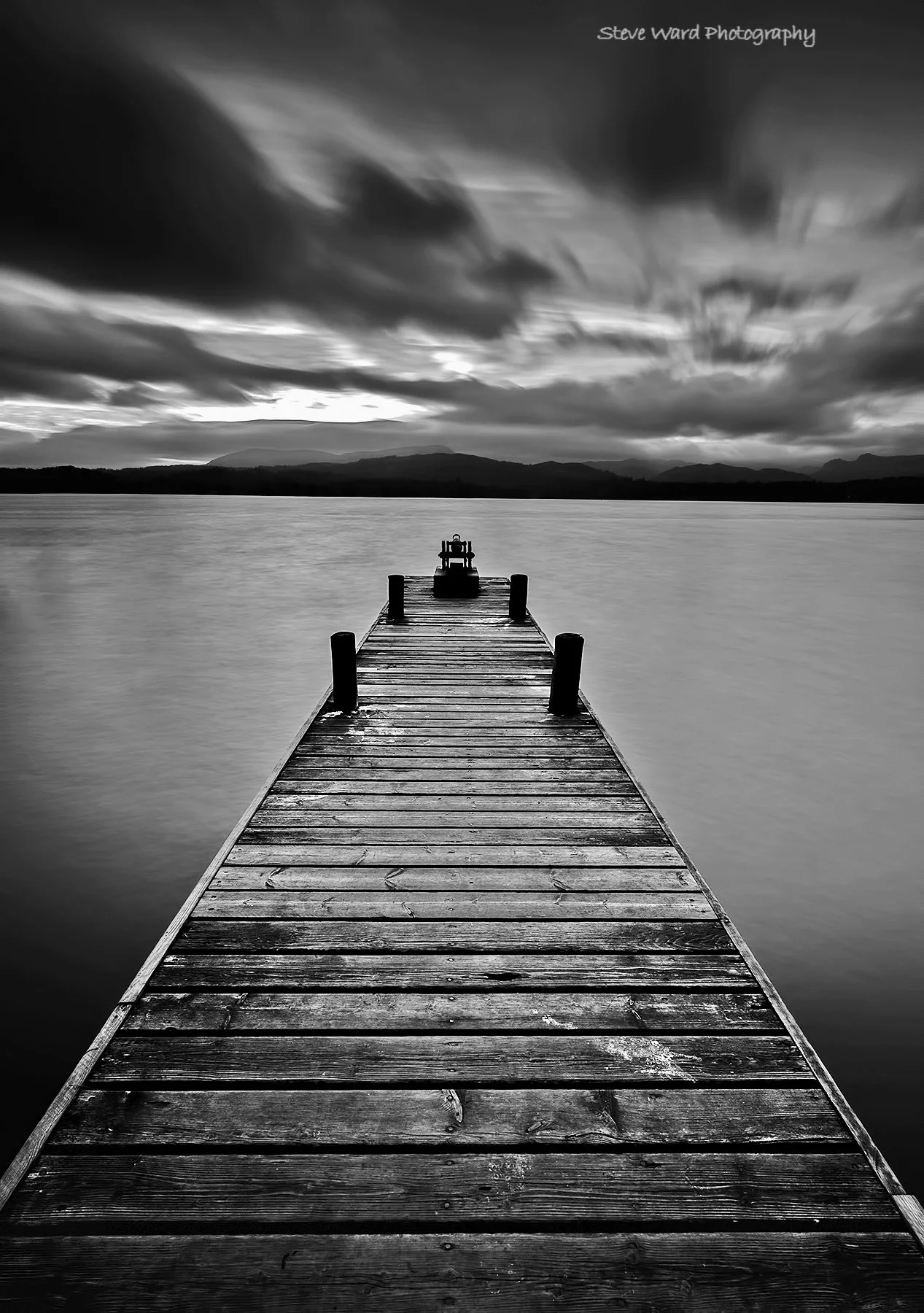 A black and white photo of a wooden dock extending into a calm body of water with mountains and a cloudy sky in the background.