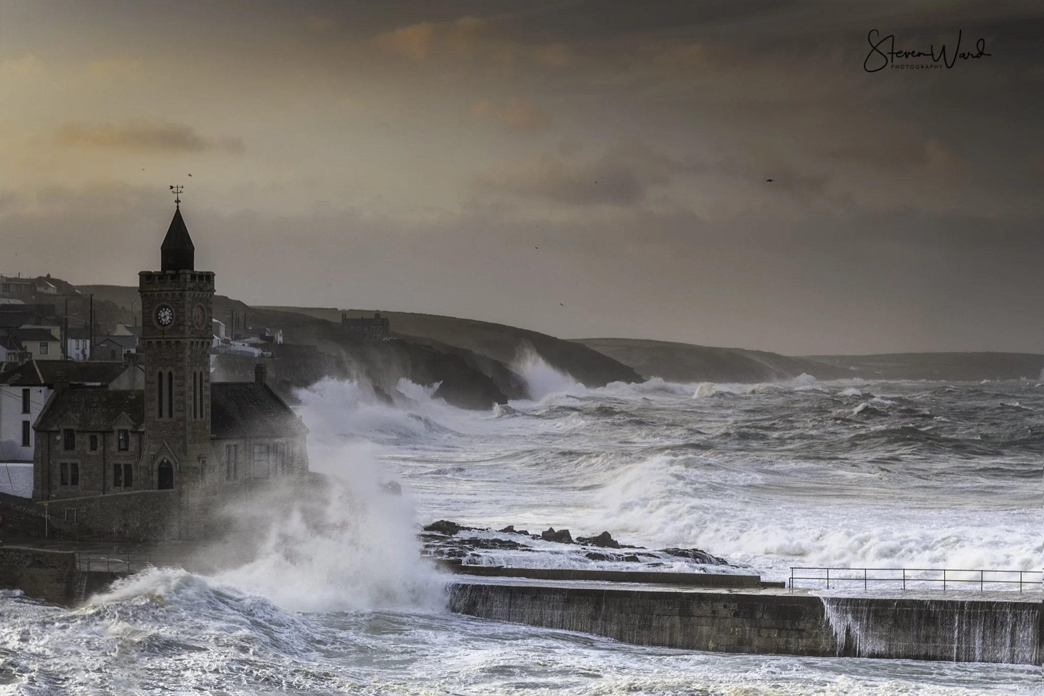 Stormy seas crashing against a lighthouse and coastal town at sunset.