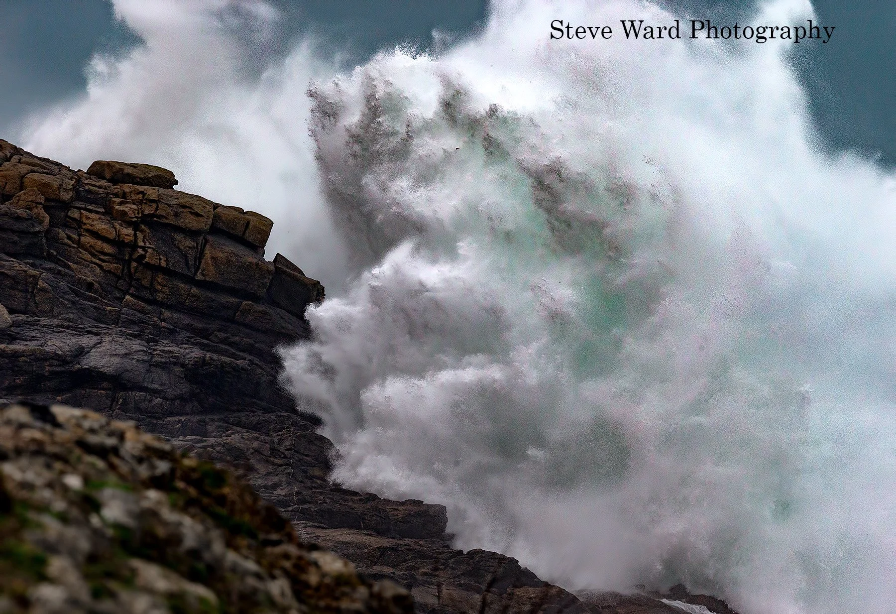 Waves crashing against rocky shoreline during a storm, with white spray and dark clouds in the background.