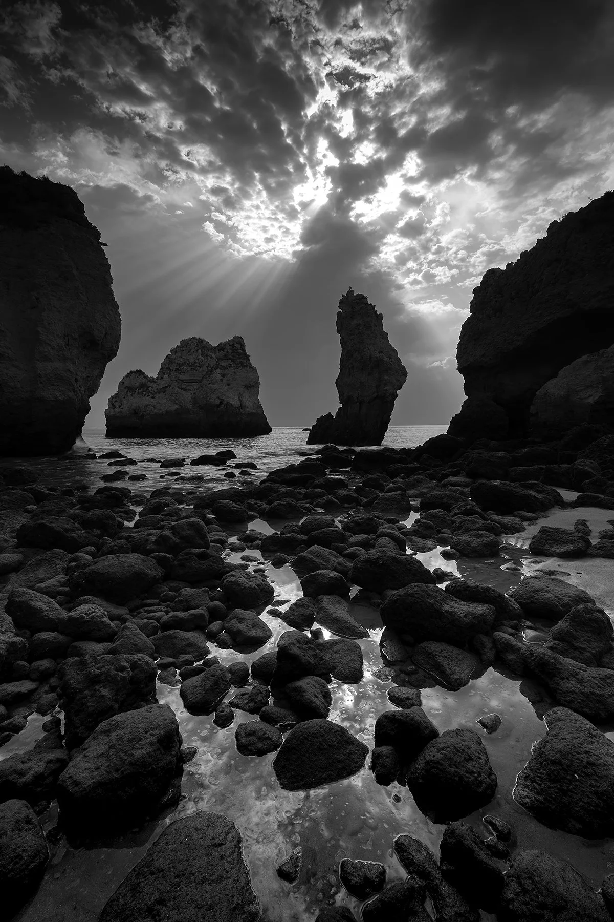 Black and white photo of a rocky beach with large rocks and sea stacks, with the sun partly obscured by clouds and rays of light shining through.
