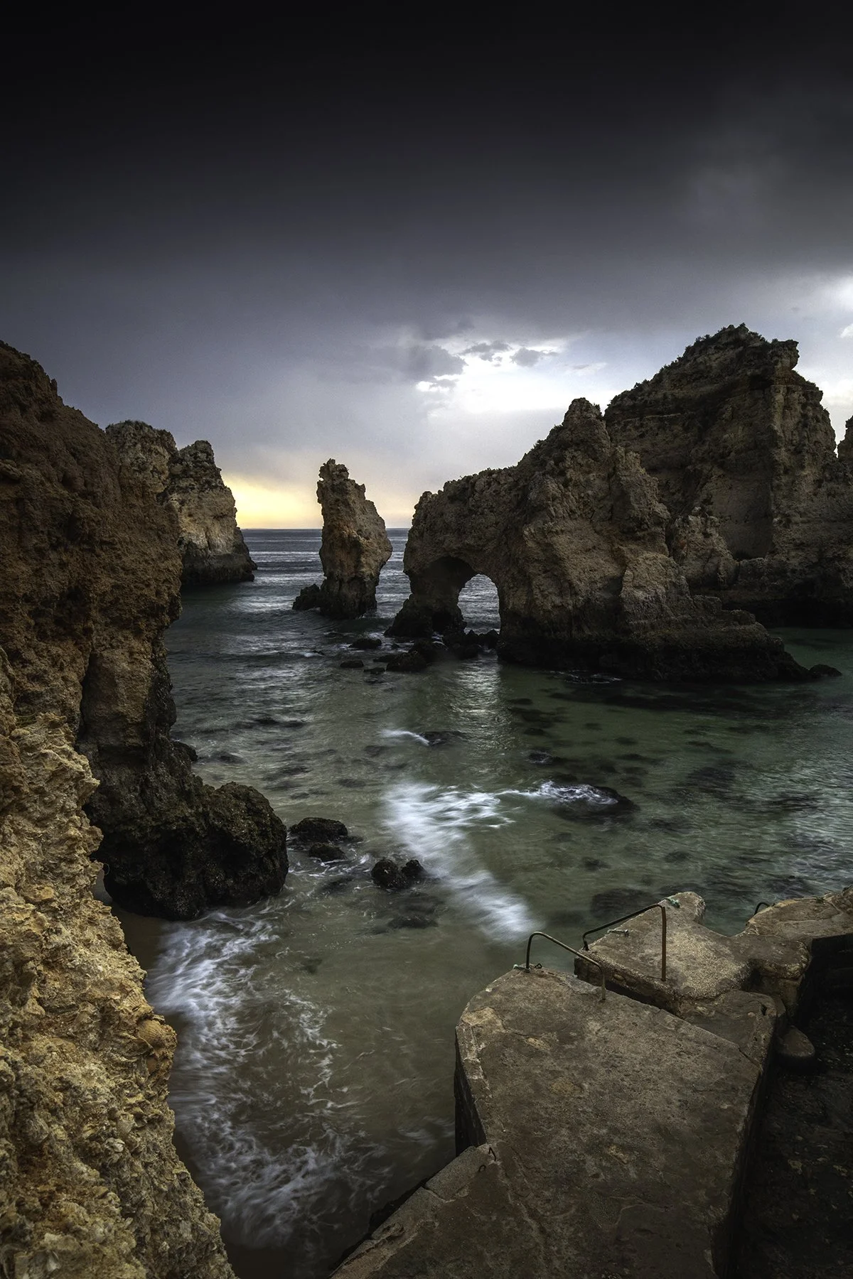 Rock formations and sea stacks at the coast during sunset with cloudy sky, waves crashing on the shore, and a concrete platform with metal railings in the foreground.