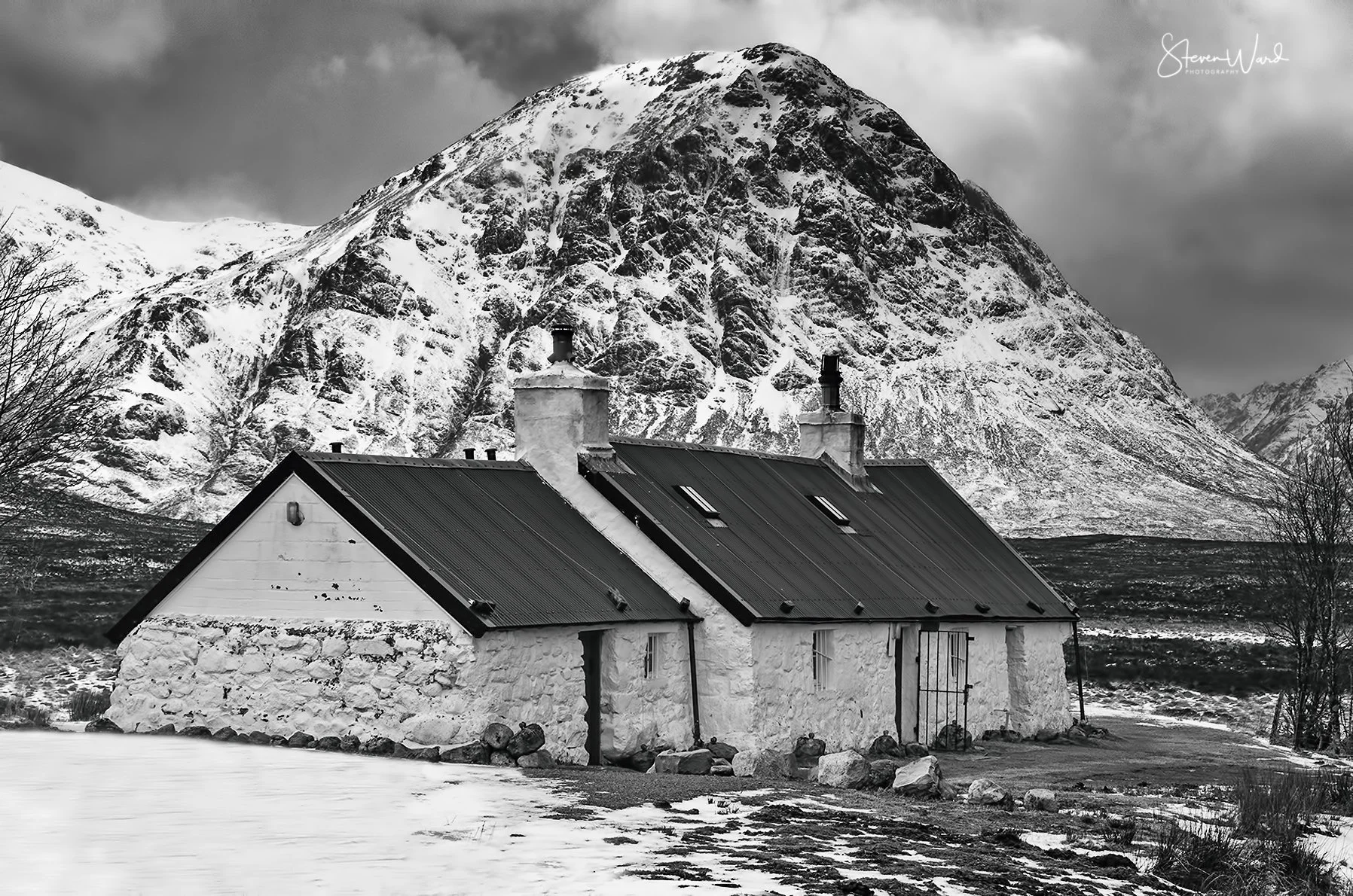 Black and white photo of a rustic house with a metal roof, chimney, and stone foundation set in a snowy landscape, with a large, snow-covered mountain in the background.