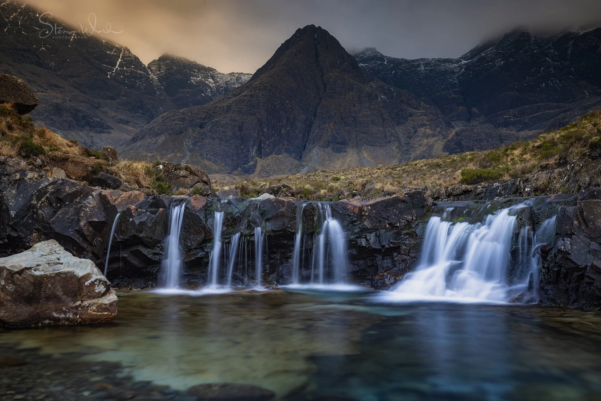 A mountain landscape with a waterfall flowing over rocks into a shallow pool, surrounded by rugged terrain and towering peaks, under a cloudy sky.
