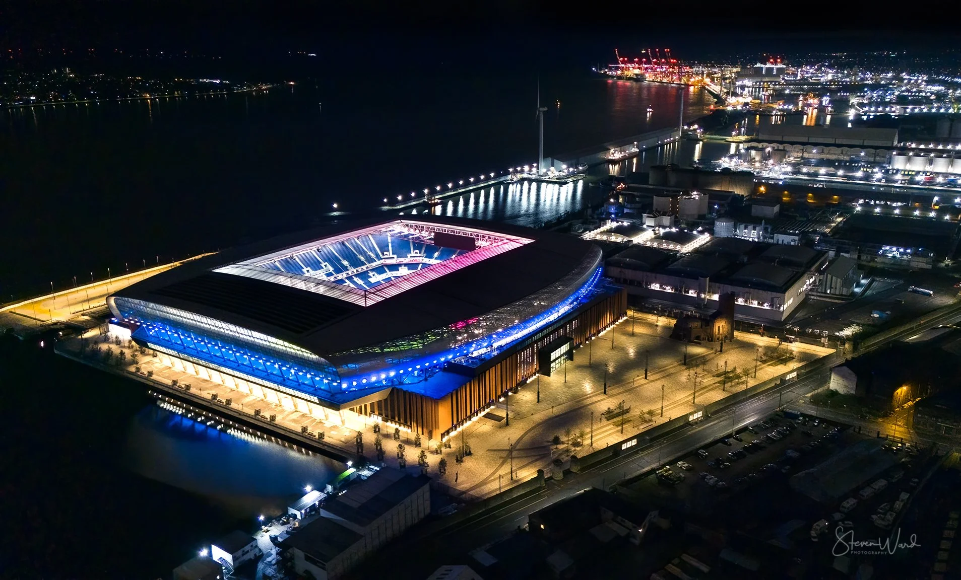Night aerial view of a sports stadium illuminated with blue and pink lights, situated by water with reflections, surrounded by industrial and port area with cranes and ships in the distance.