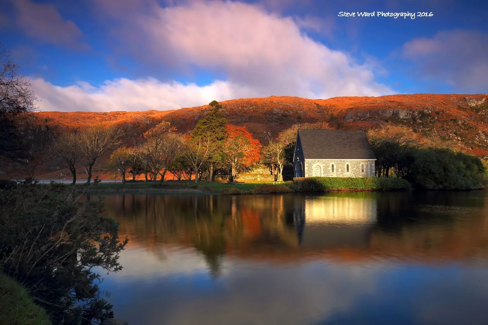 A scenic landscape featuring a small stone church with a dark roof, situated near a reflective body of water. The background shows a mountain with reddish-brown foliage, trees with sparse leaves, and a partly cloudy sky with patches of blue and soft 
