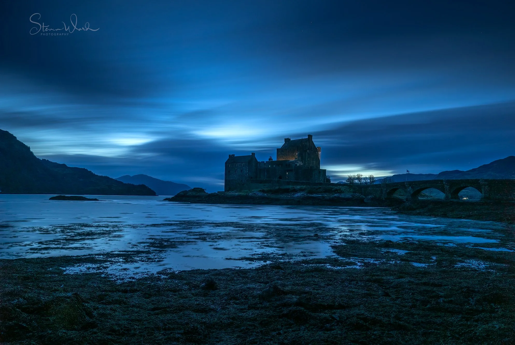 A castle on a small island surrounded by water, with a stone bridge leading to it, under a dark blue sky with clouds, mountains in the background.