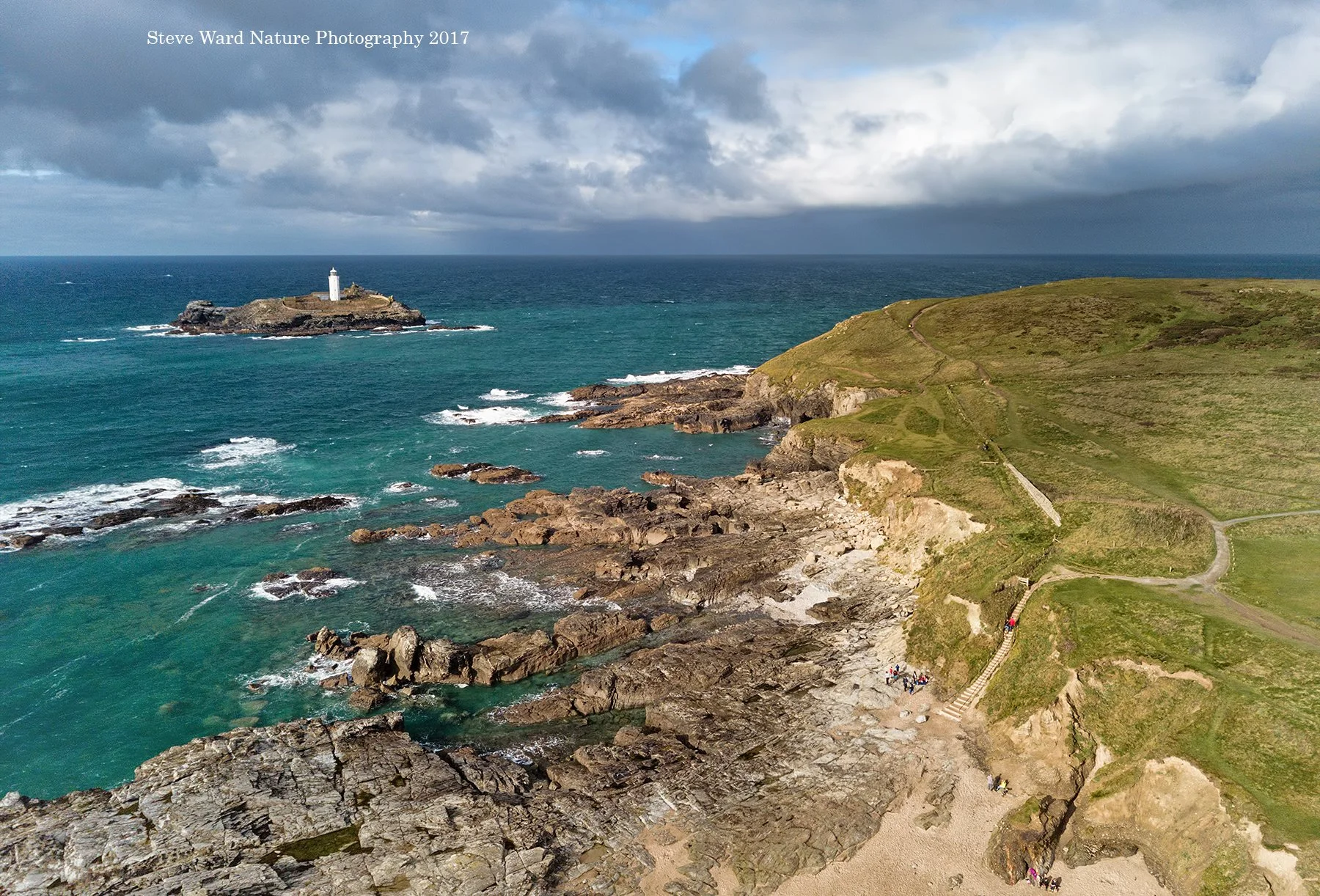 A rugged coastline with rocky shorelines and green grassy hills. A lighthouse stands on a small island in the ocean. A group of people walk along a path on the hill, and the sea has waves with whitecaps, under a partly cloudy sky.