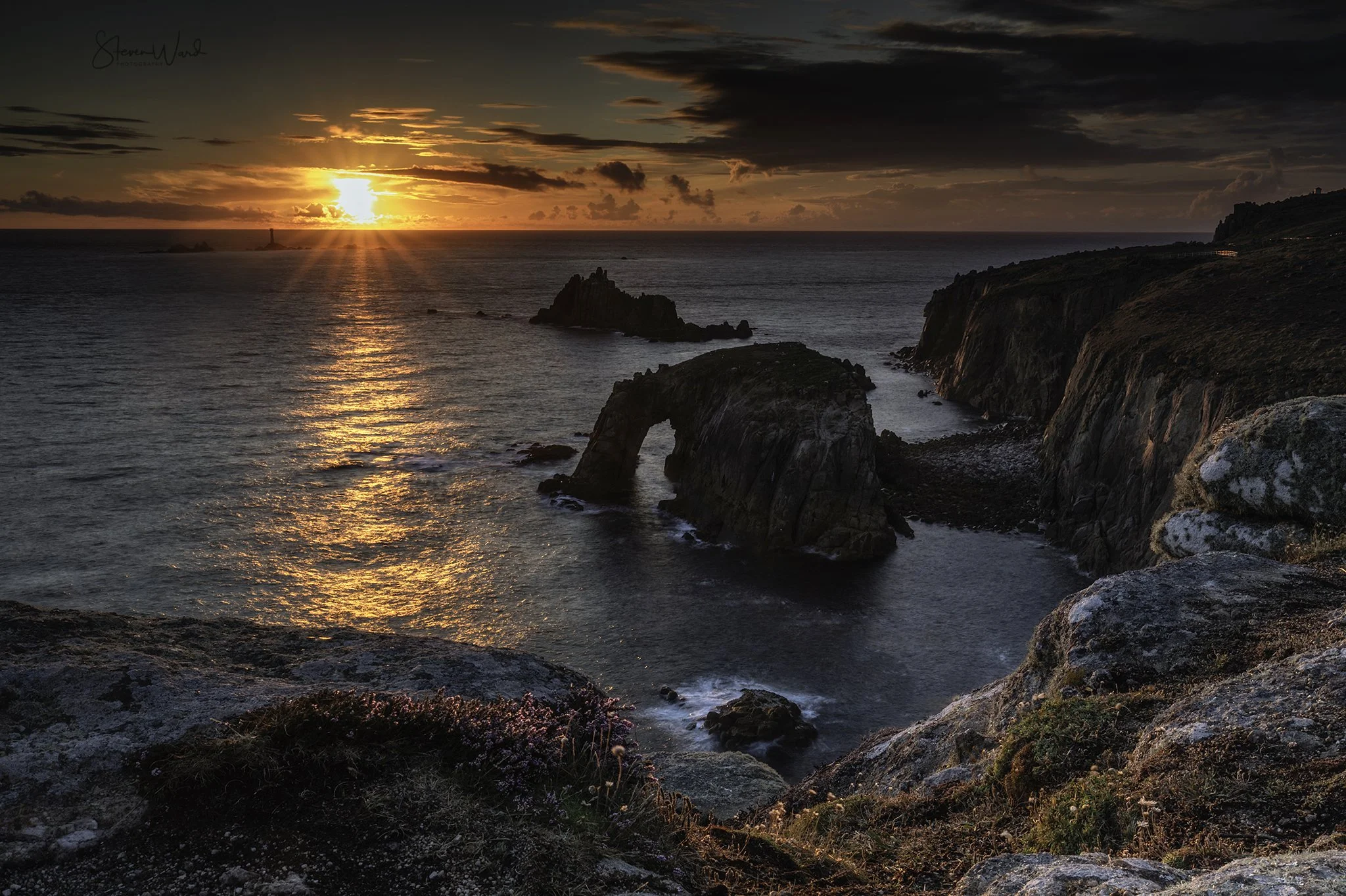 A coastal landscape at sunset with rocky cliffs, a natural rock arch formation in the water, and the sun setting on the horizon reflecting orange and yellow colors on the ocean surface.