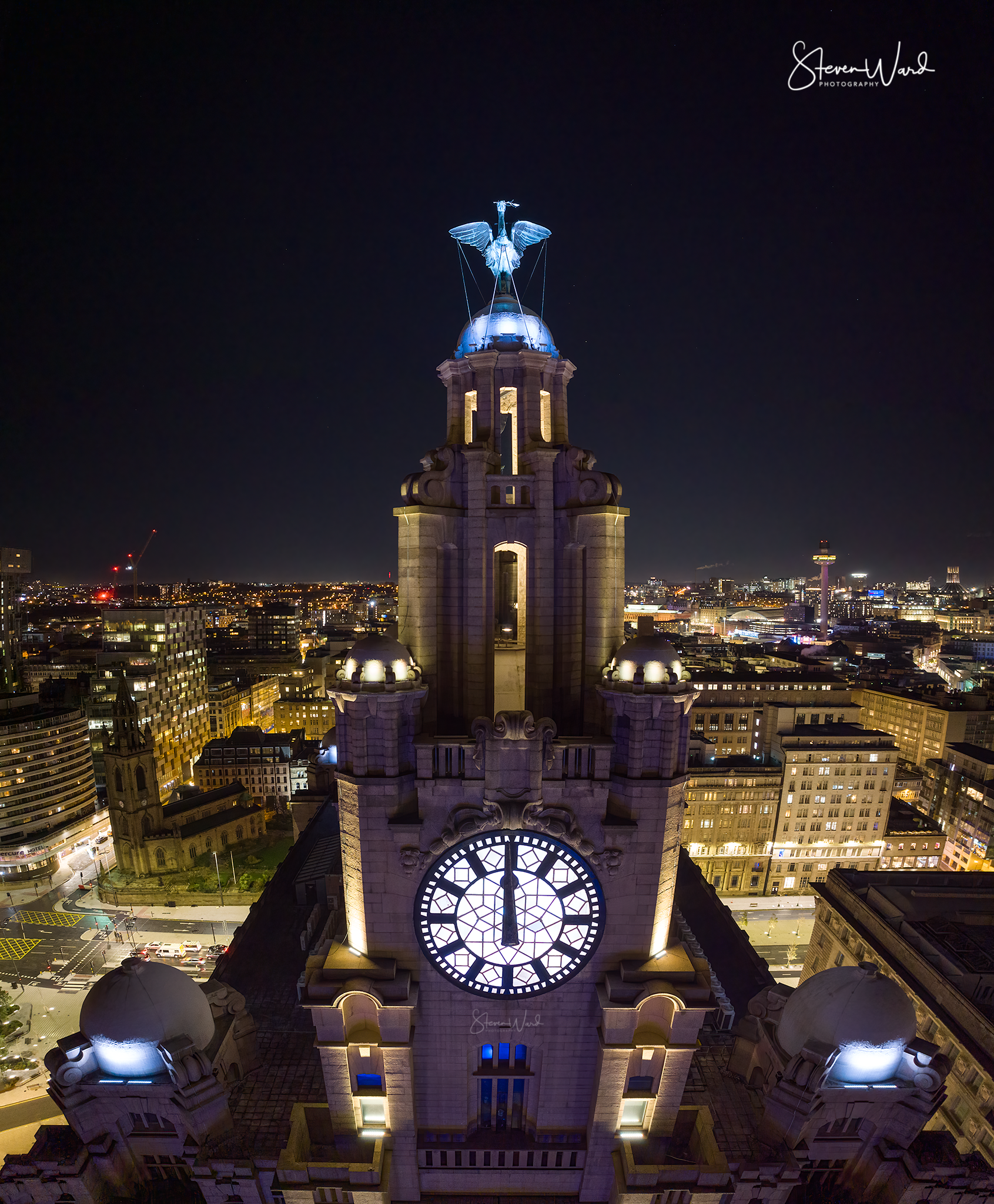 Nighttime aerial view of a historic clock tower with ornate architecture, illuminated at night, with city lights and skyline in the background.