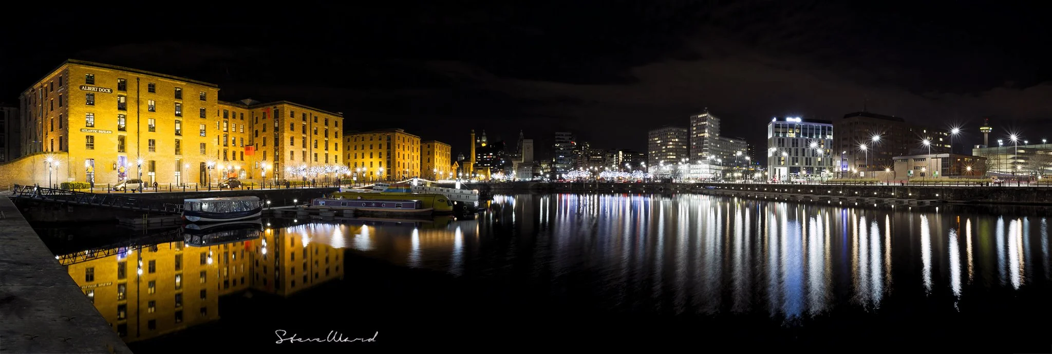 Night view of a city marina with brightly lit buildings and reflections on calm water.
