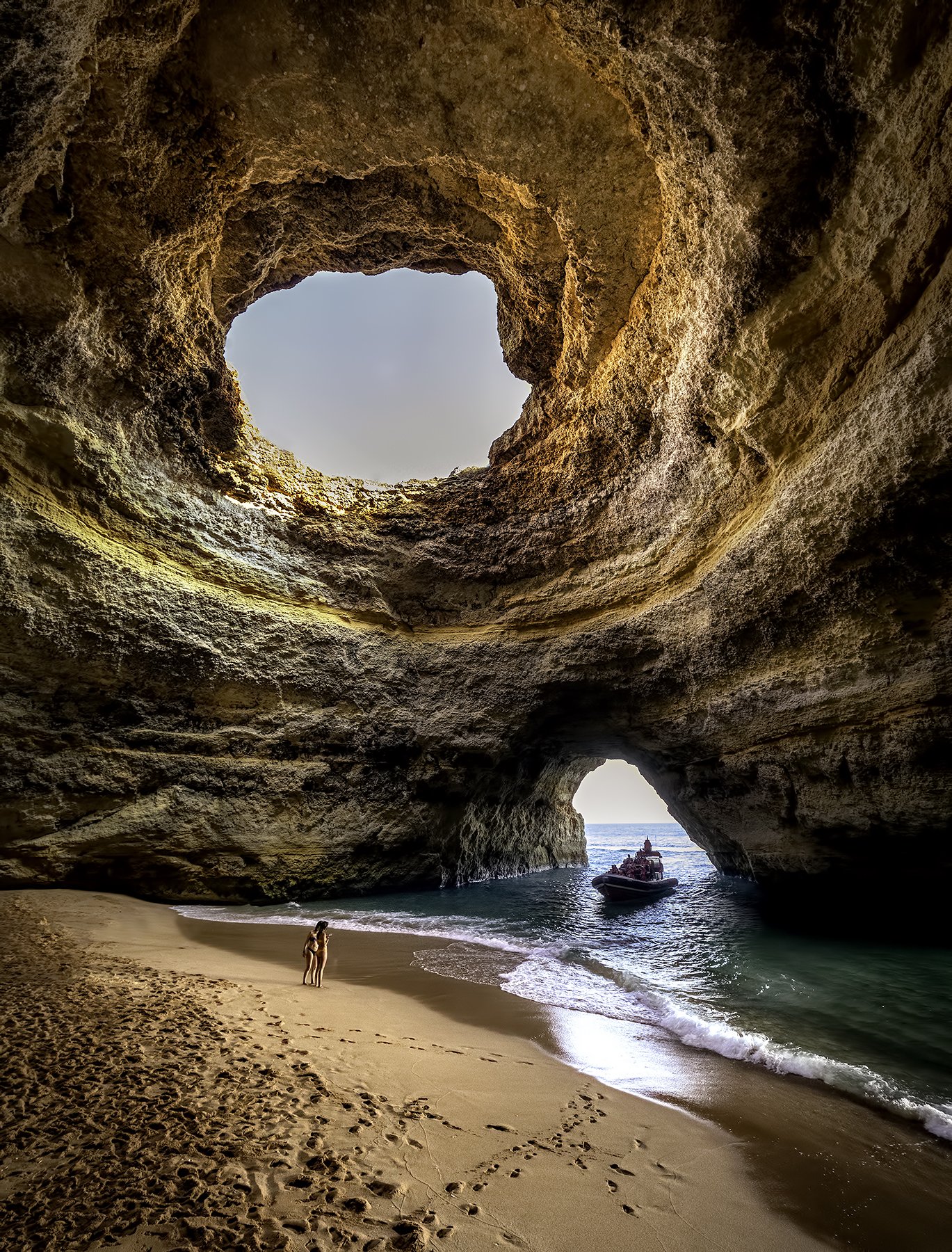 Inside a seaside cave with large opening, sandy beach, and a boat in water. Woman standing on the beach.