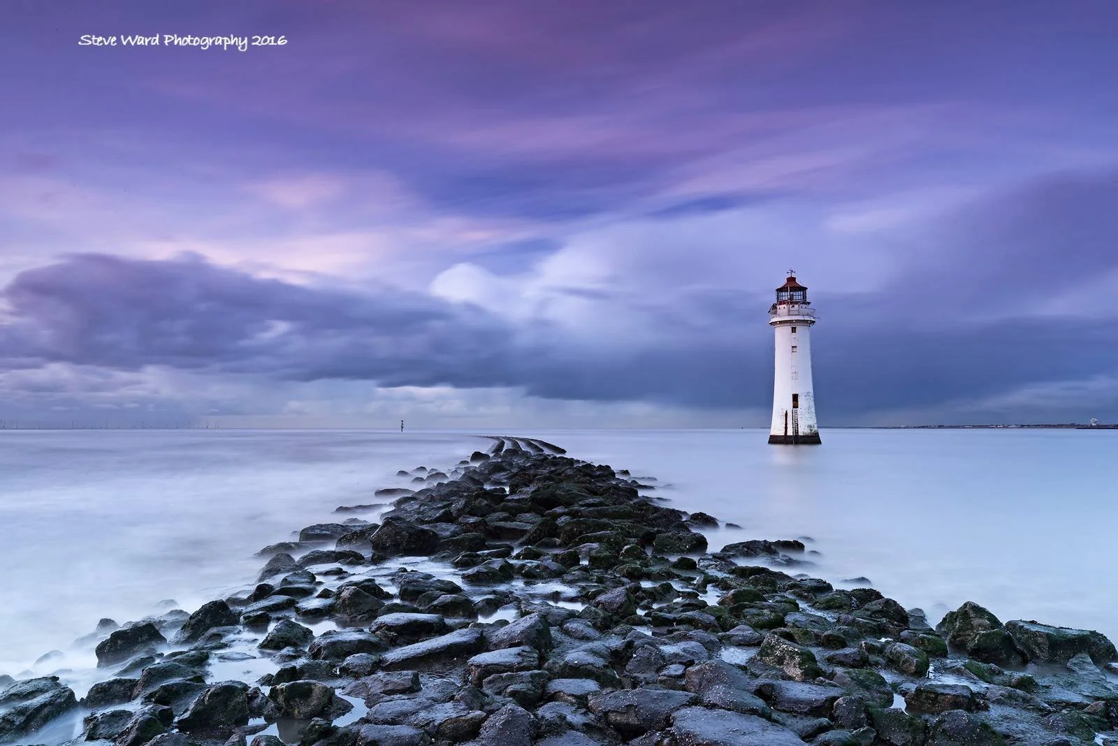 A lighthouse on a rocky breakwater extending into calm water, with a cloudy sky and purple hues.