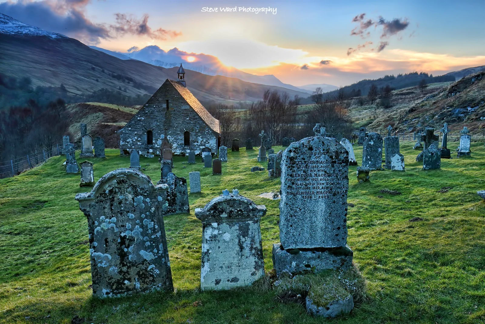 A historic graveyard with weathered tombstones in front of a stone church, mountains in the background, and a sunset sky.