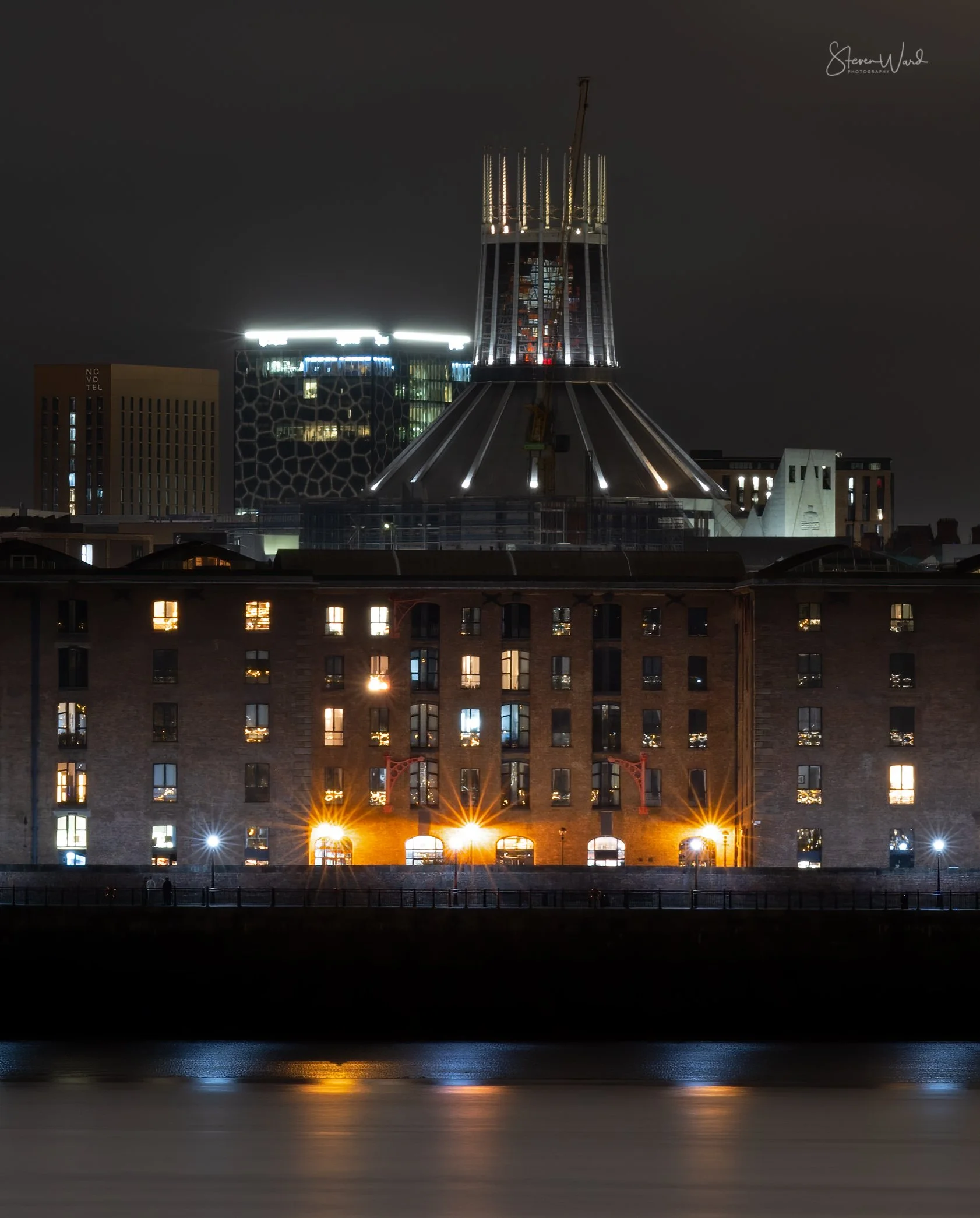 Nighttime cityscape with illuminated buildings, reflecting water, and a construction crane on a tall building under construction.